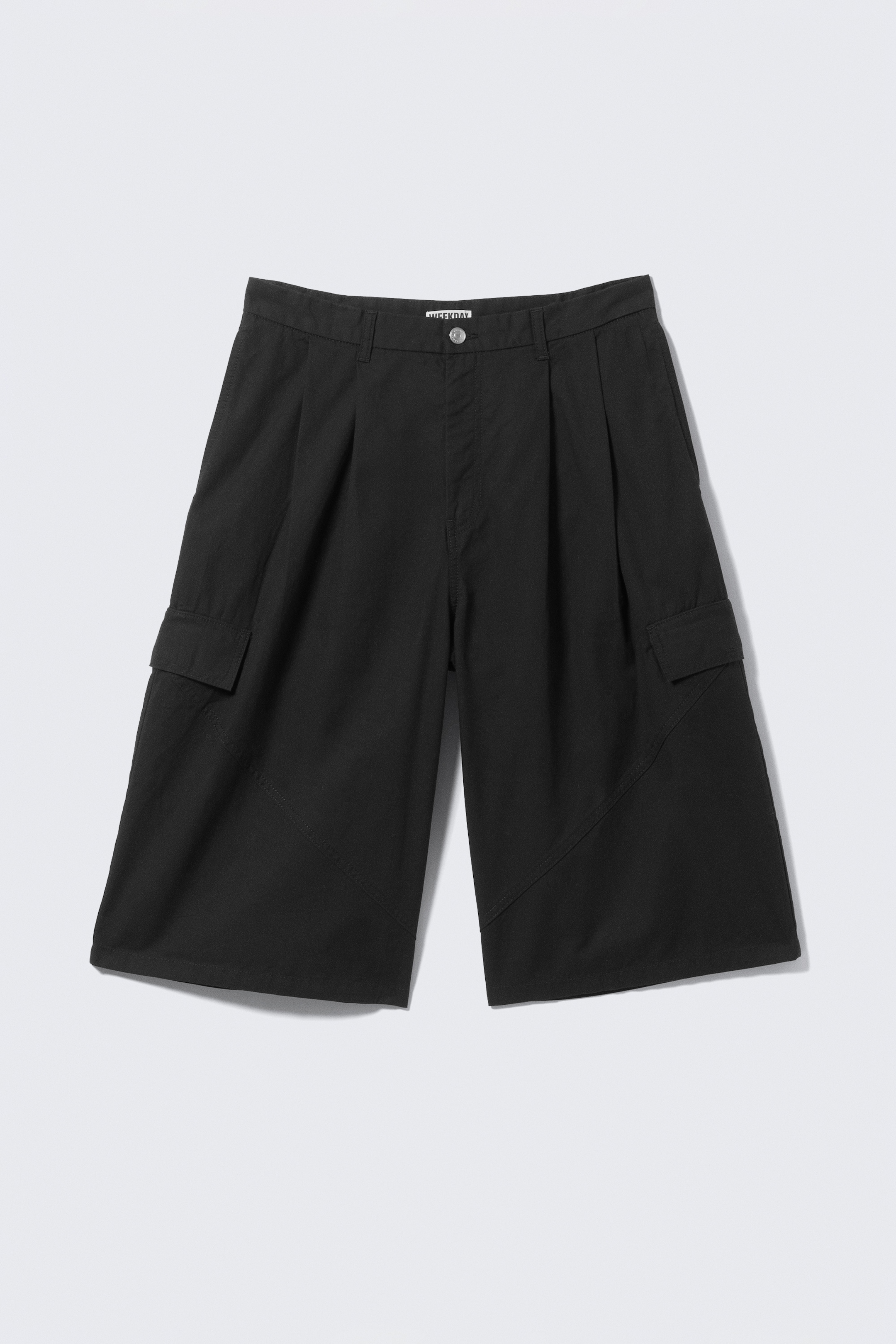 Größeres Bild ansehen: Ein Paar schwarze, locker sitzende, knielange Shorts wird von vorne präsentiert. Gefertigt aus Baumwolltwill, zeichnen sich die Shorts durch vordere Falten, abgeschrägte Seitennähte und Klappentaschen an den Oberschenkeln aus. Gürtelschlaufen und ein Reißverschluss mit Knopfverschluss vervollständigen die strukturierte Taille.