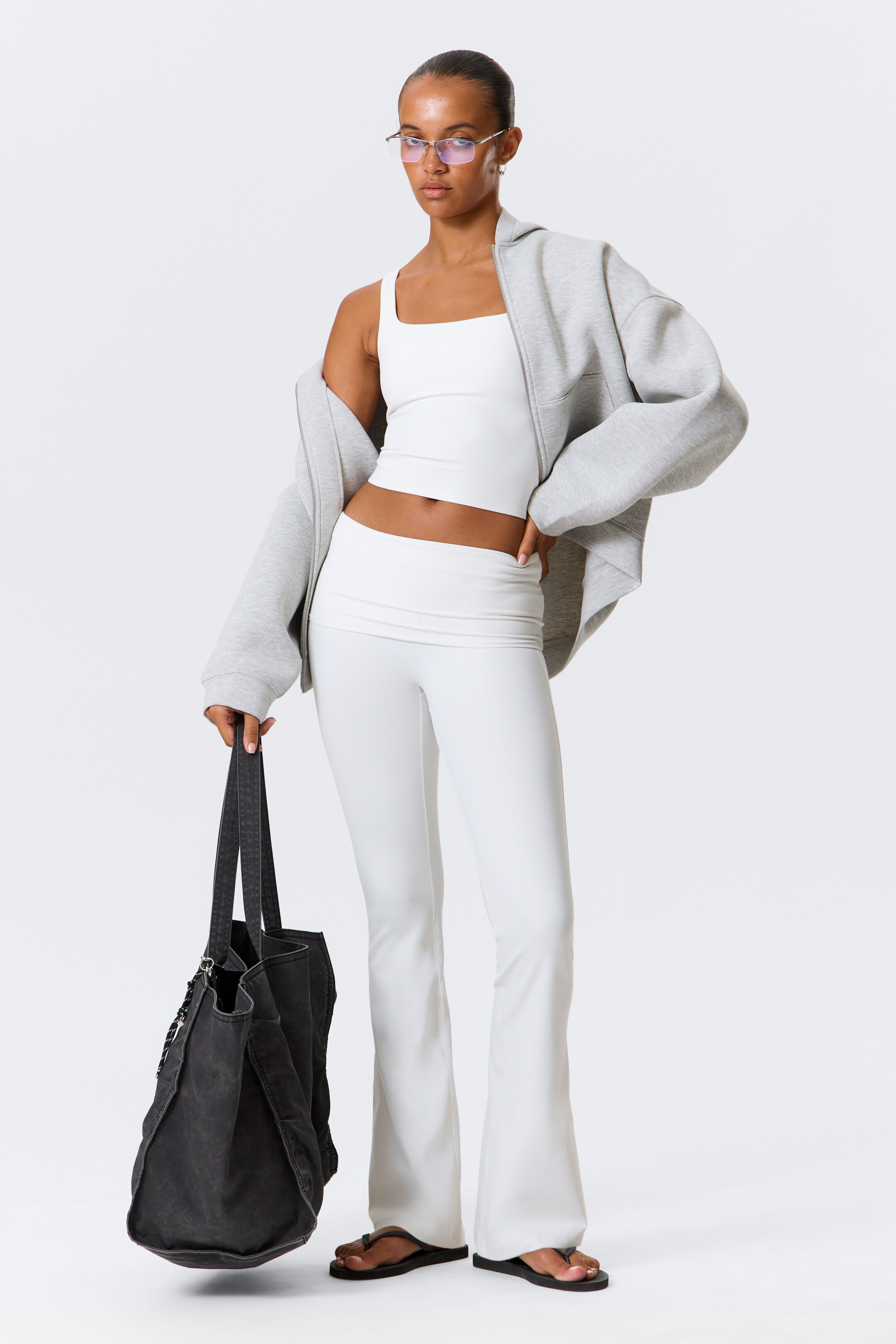 Agrandir l'image: Un sweat à capuche zippé surdimensionné gris clair est porté ouvert sur un débardeur court à encolure carrée blanc et un pantalon de yoga évasé blanc avec une large ceinture repliée. L'ensemble est stylisé avec des tongs noires, un sac fourre-tout en toile gris foncé usé et des lunettes sans monture, tel que présenté sur un mannequin légèrement tourné vers la droite.