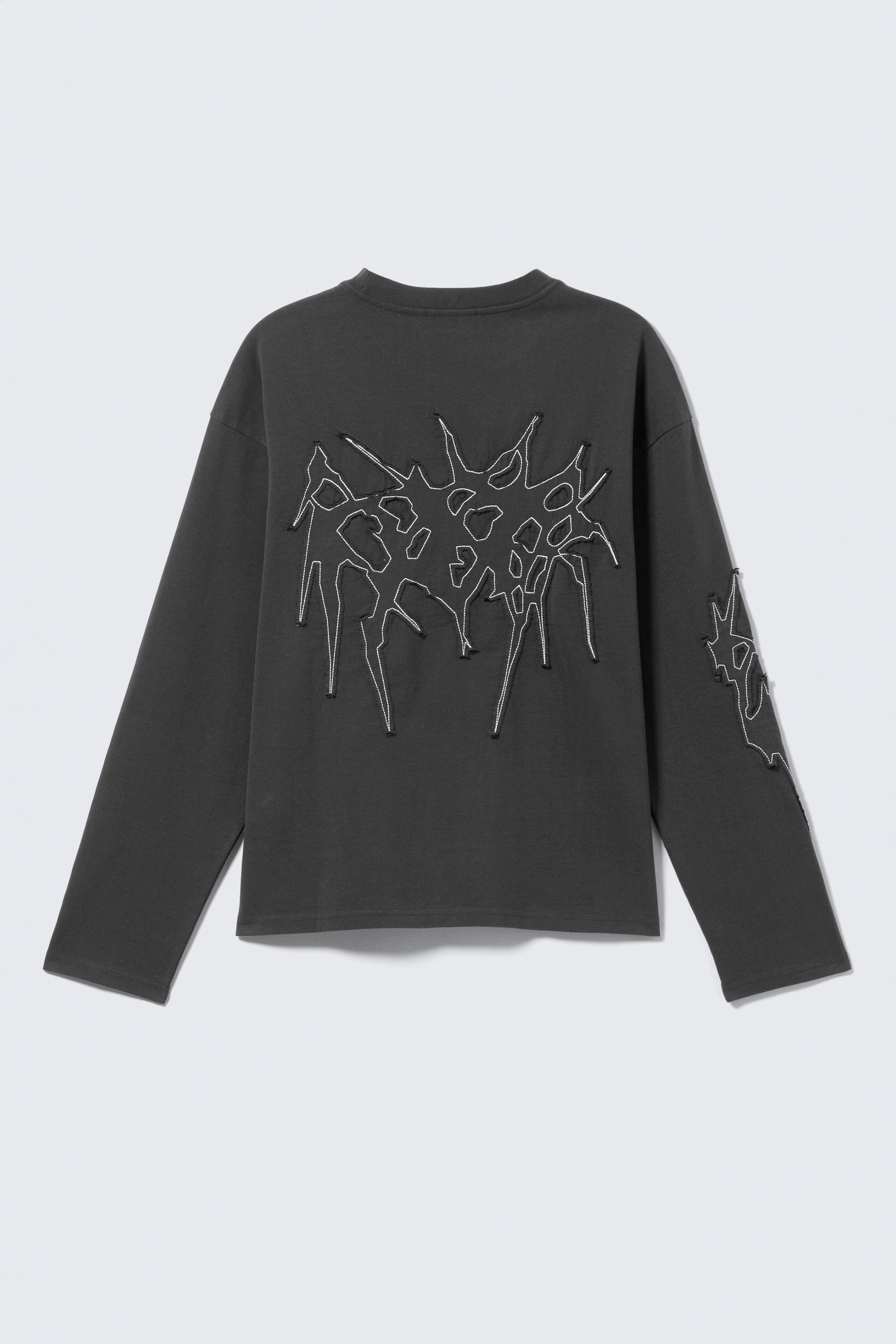 Zobraziť väčší obrázok: Tmavosivá tričko s dlhými rukávmi predstavuje svoj CHRBÁT, s okrúhlym rebrovaným výstrihom a boxovým pravidelným strihom. Na chrbte a na hornej časti ľavého rukáva sa nachádza veľký, abstraktný, nepravidelný dizajn, ktorý je orámovaný bielym prešívaním. Dvojité šitie je viditeľné na manžetách a spodnom leme.