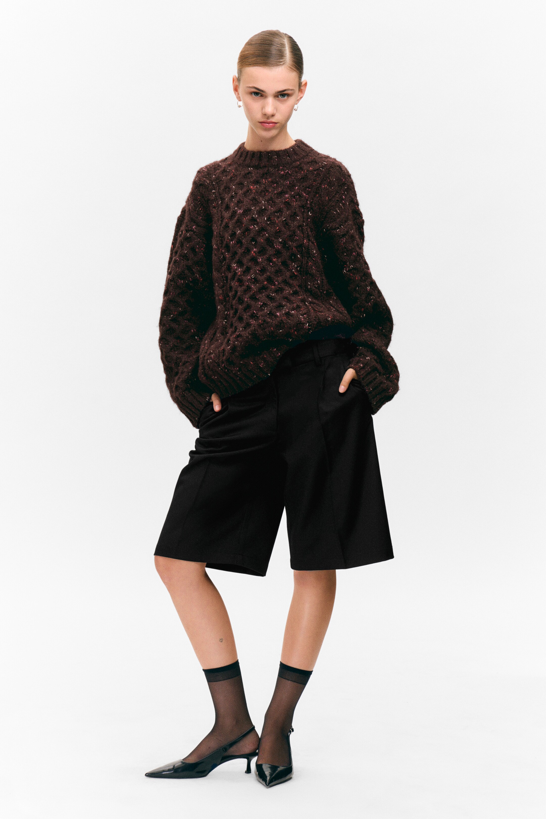 Se større billede: En mørkebrun chunky strikket sweater med et åbent vævemønster og plettet tekstur præsenteres fra en let vinkel, der viser dens oversized pasform og voluminøse ærmer. Den er parret med sorte skræddersyede Bermuda-shorts, som viser et afslappet, knælængde snit og strukturerede frontsømme.