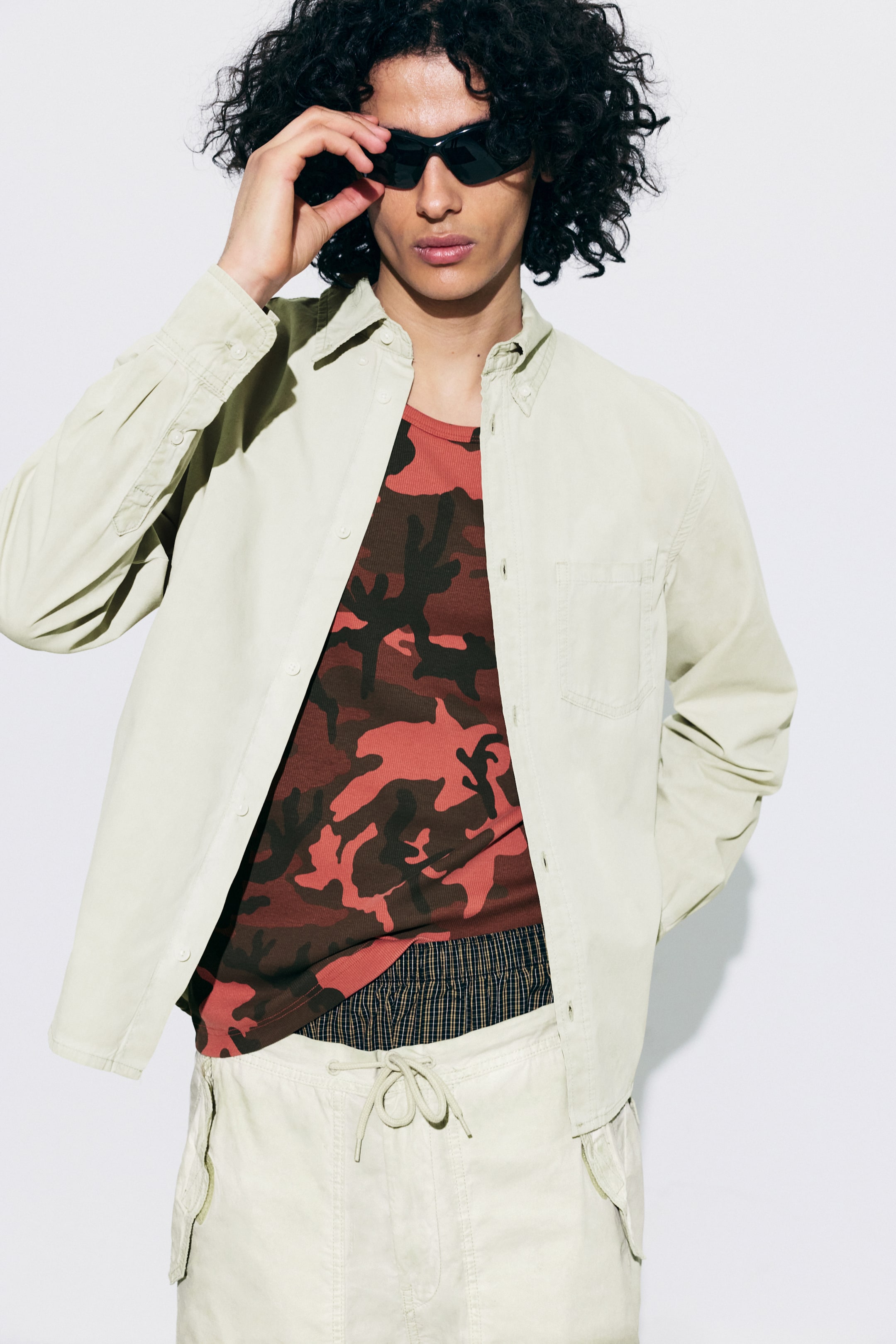 Grotere afbeelding bekijken: Een licht beige overhemd met knopen wordt open gedragen, waardoor een rood camouflage t-shirt zichtbaar is dat over geruite boxers is gelaagd. Licht beige cargoshorts met een zichtbare trekkoord taille worden ook gedragen, samen met zwarte cat-eye zonnebrillen die worden vastgehouden.