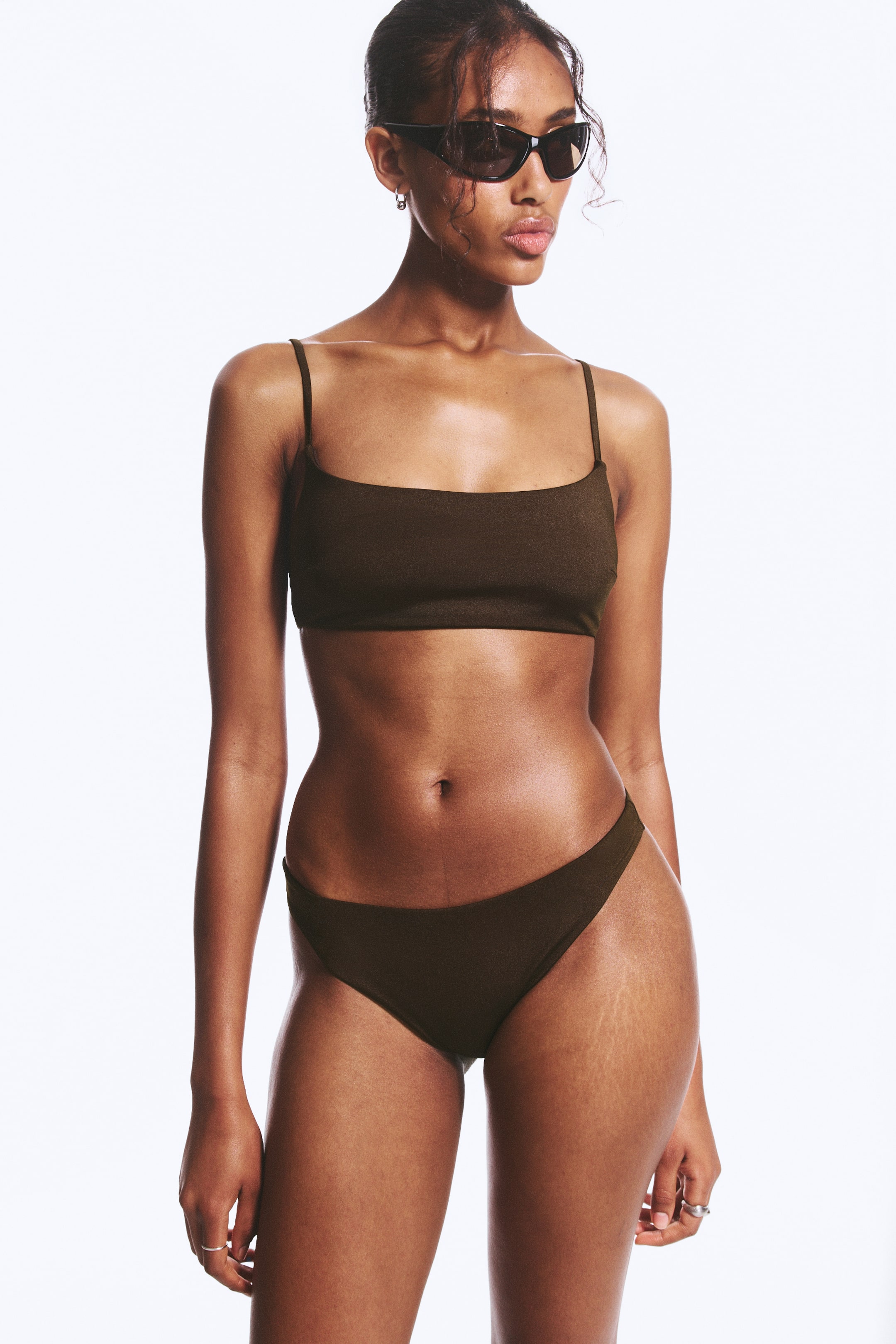 Grotere afbeelding bekijken: Een donkerbruin tweedelig badpak wordt gepresenteerd, met een bralette top met vierkante hals en dunne spaghettibandjes, en bijpassende hoog uitgesneden bikinibroekjes. De stof heeft een subtiele textuurafwerking.