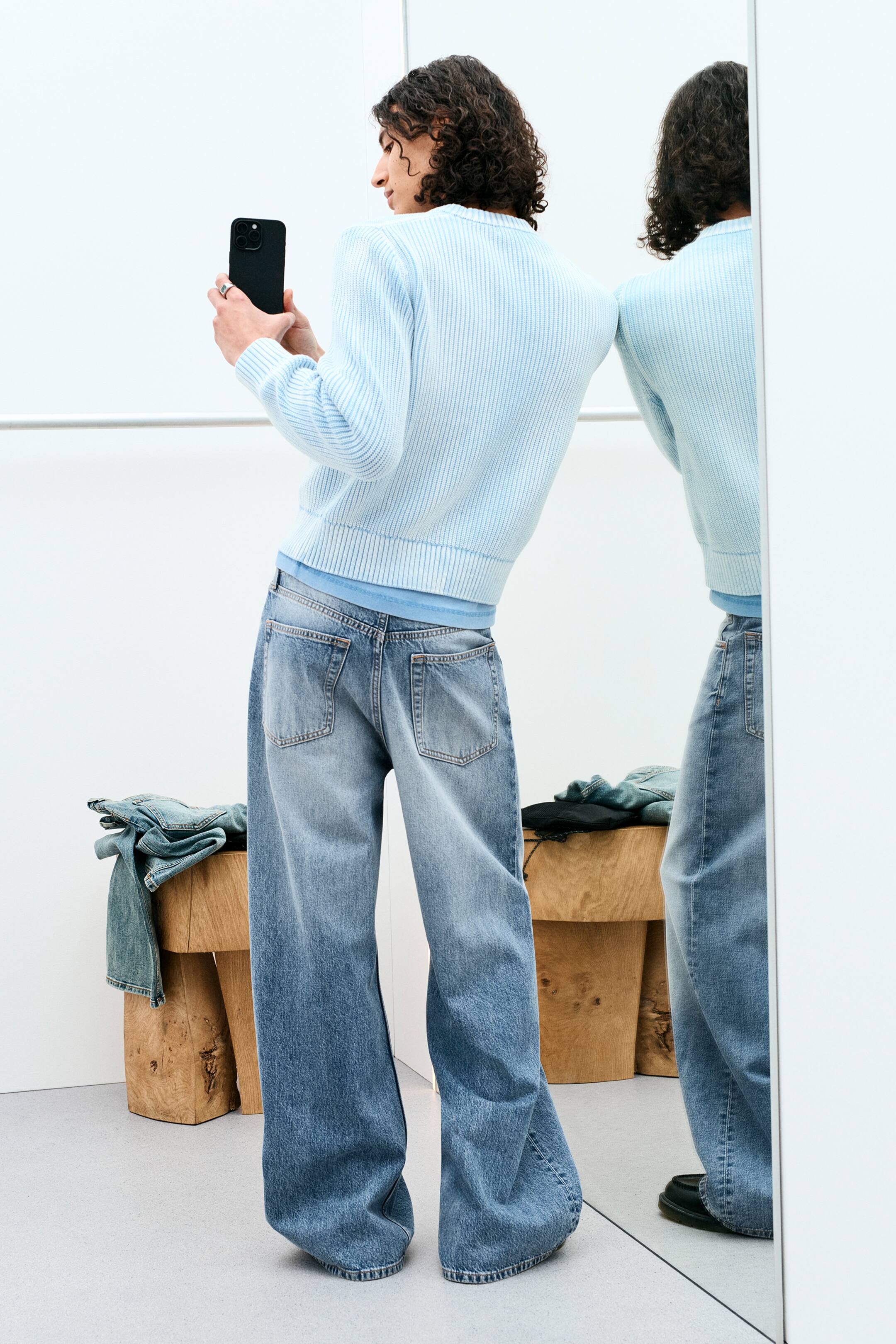 Größeres Bild ansehen: Ein hellblauer, gerippter Rundhalspullover wird über einem helleren blauen T-Shirt getragen und mit locker sitzenden, hellen Denim-Jeans kombiniert, die sichtbare Gesäßtaschen aufweisen. Das Outfit wird von hinten und seitlich präsentiert, leicht nach rechts gewinkelt, um den entspannten Sitz und das weite Bein-Design zu zeigen.