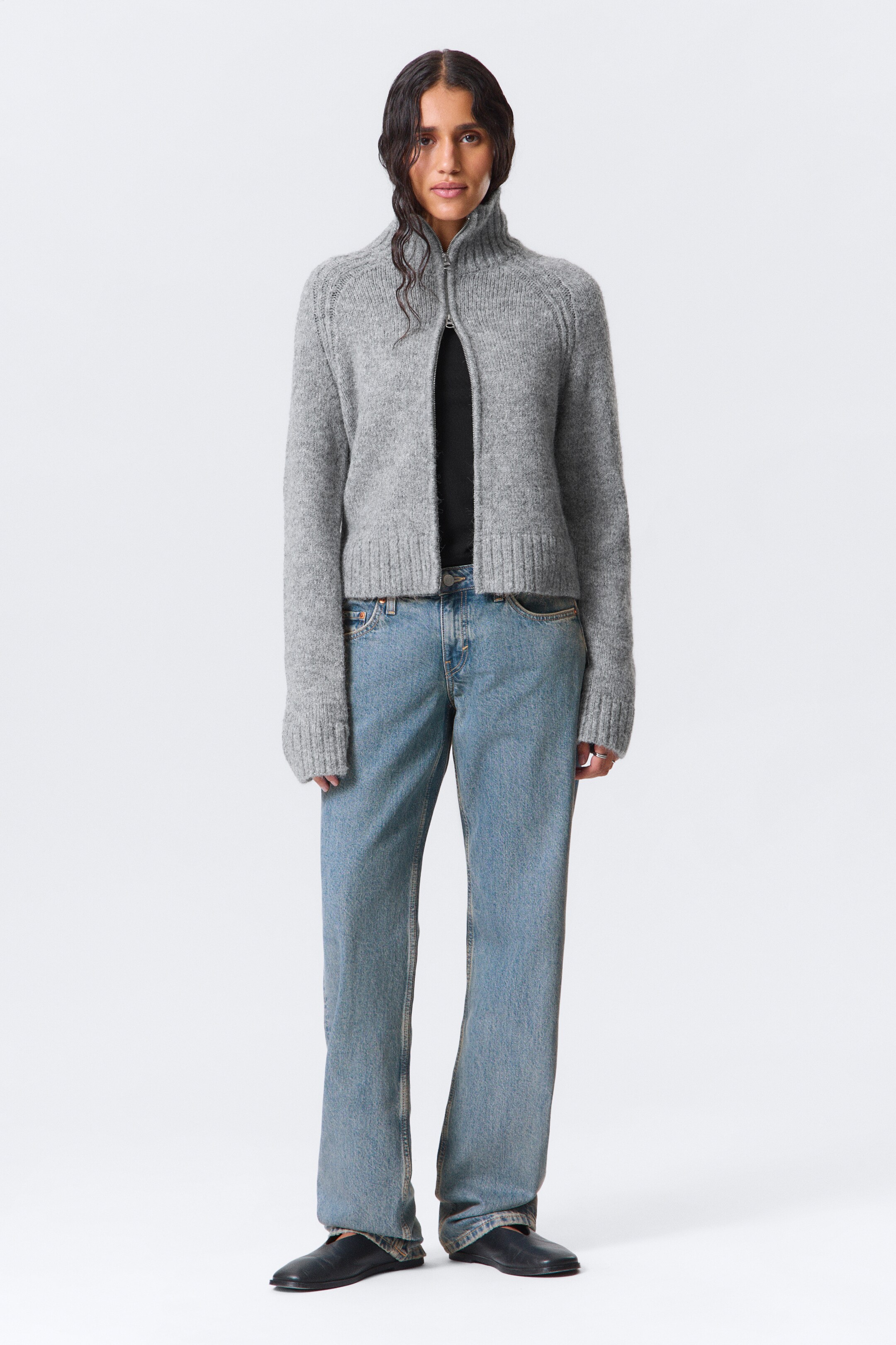 Agrandir l'image: Un cardigan gris à fermeture éclair intégrale en maille est porté, face avant. Il présente une longueur courte, un col montant, des manches raglan et des poignets et un ourlet côtelés, avec une fermeture éclair de couleur argent partiellement ouverte sur un haut noir.