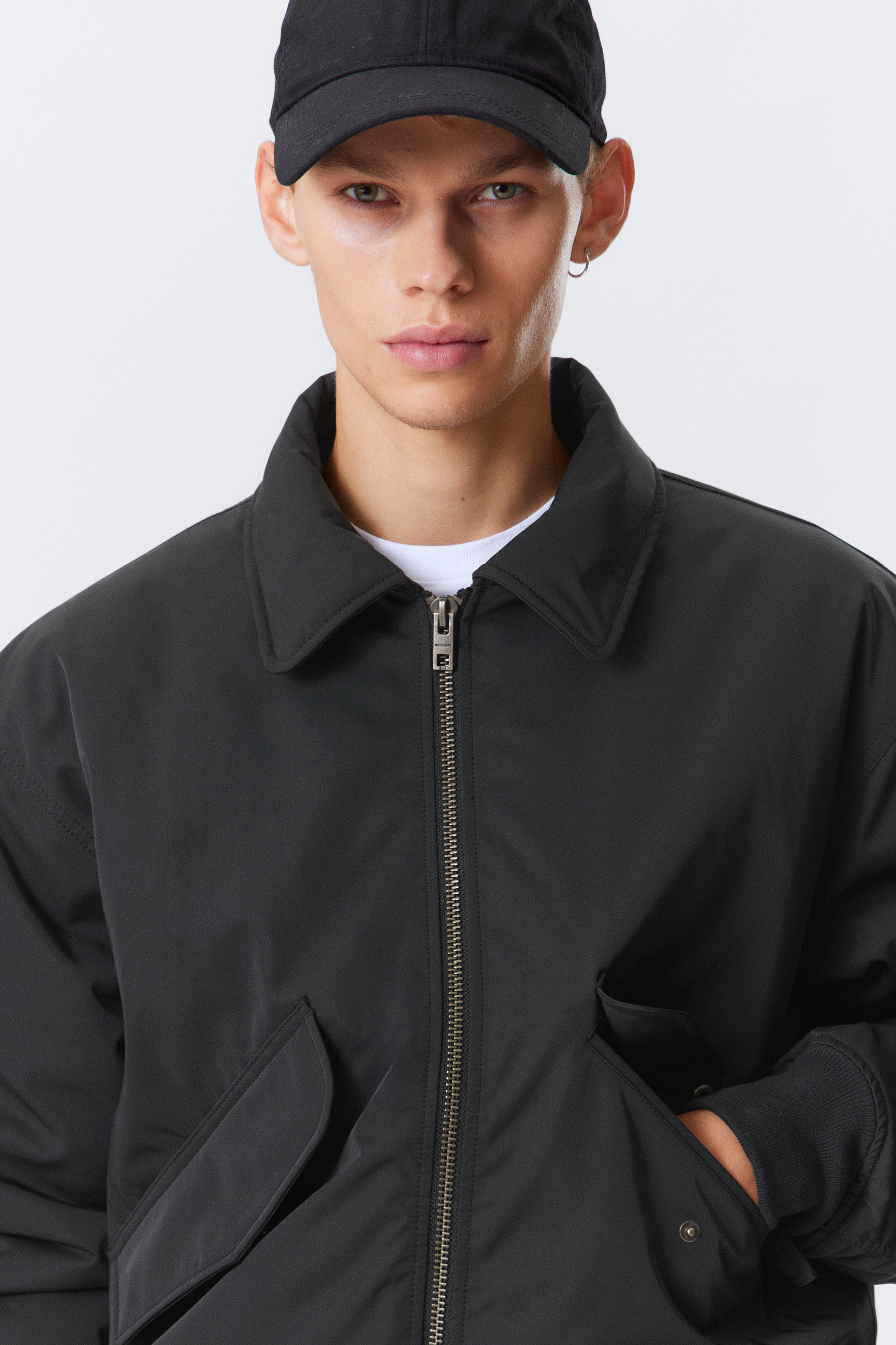Visualizza immagine più grande: Una giacca bomber imbottita nera presenta un colletto a punta, una chiusura centrale con zip metallica, tasche frontali a patta angolate e polsini a coste. La modella indossa la giacca sopra una t-shirt bianca, con una mano all'interno di una tasca frontale aperta.