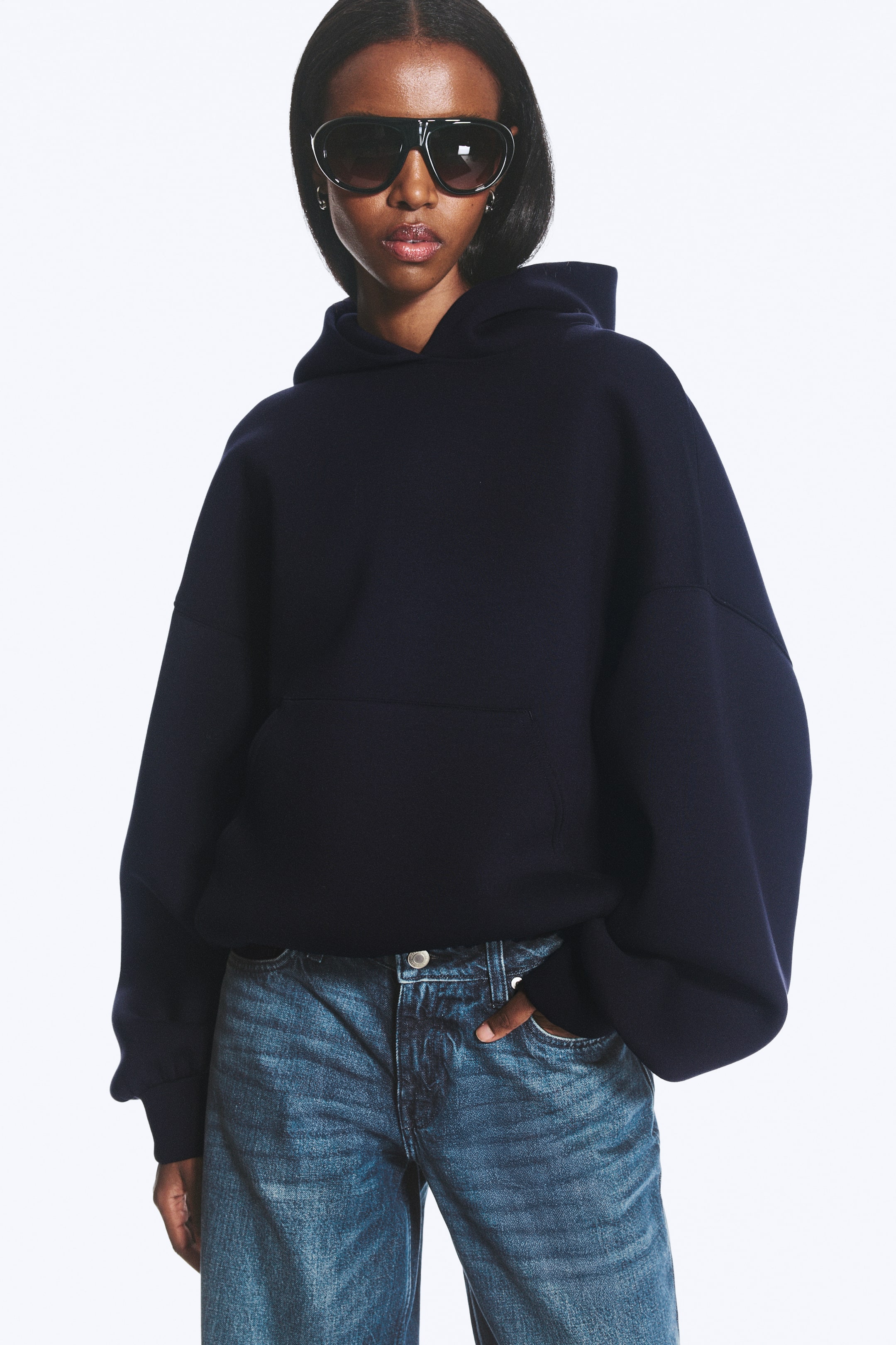 Größeres Bild anzeigen: Ein dunkelblauer Scuba-Hoodie, im Oversized-Look geschnitten, wird vom Model getragen. Er verfügt über eine geräumige Kapuze, überschnittene Schultern, eine Kängurutasche sowie glatte Bündchen und einen glatten Saum, während die Hand des Models in der Tasche der blauen Jeans ruht, die zusammen mit dem Hoodie getragen wird.