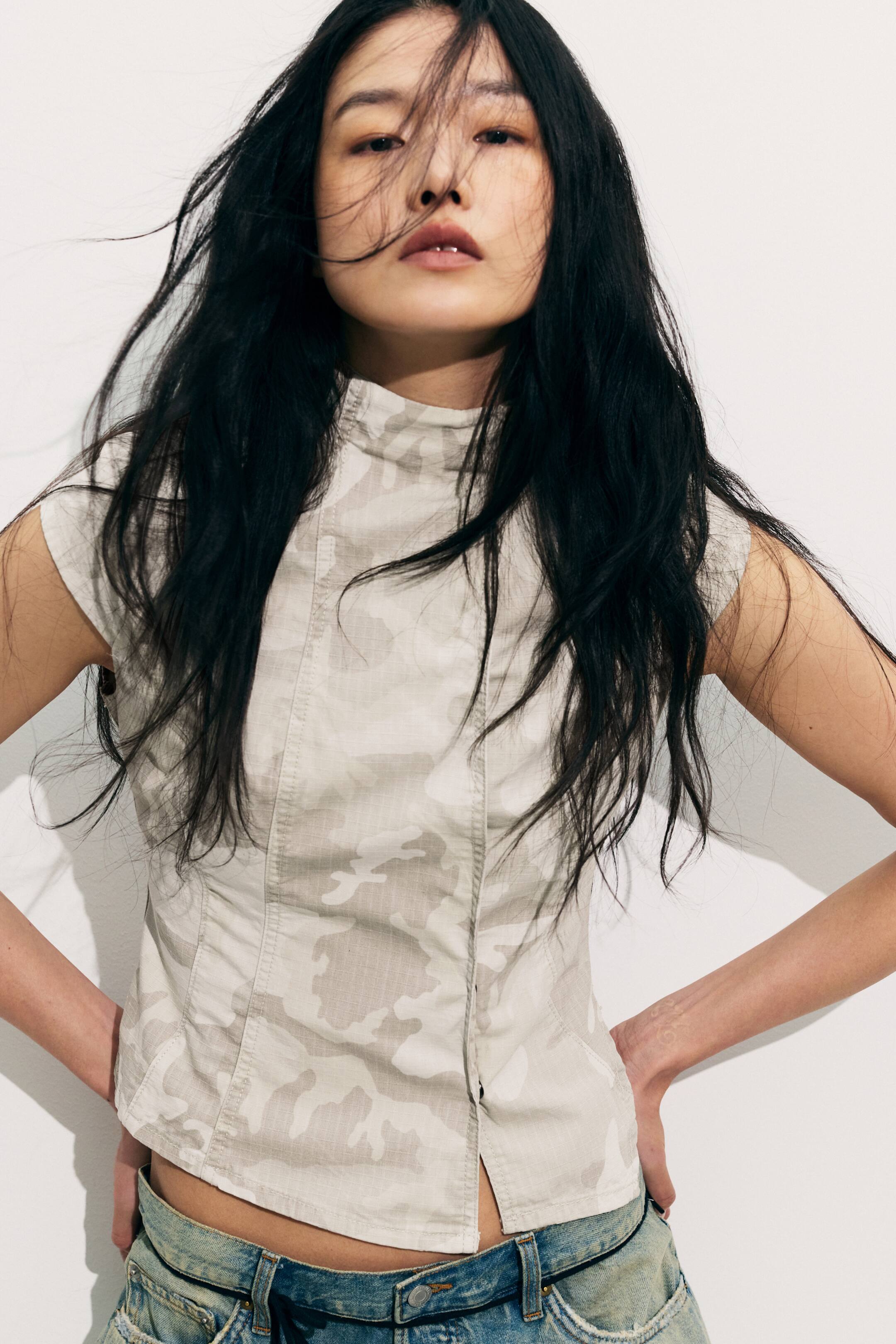 Agrandir l'image: Un haut sans manches présente un motif camouflage en gris clair et blanc cassé, un col montant et des manches courtes. Il affiche des coutures verticales à l'avant et un ourlet légèrement raccourci avec une petite fente à l'avant.