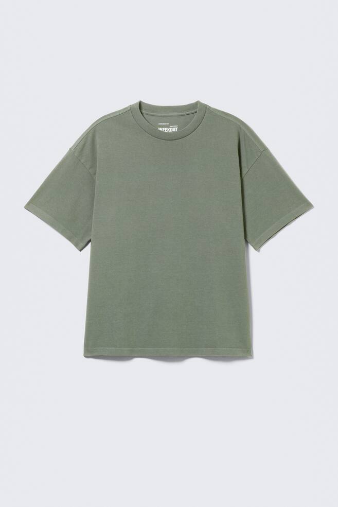 Una camiseta verde oliva se muestra de frente, con un corte holgado y suelto, hombros caídos y mangas cortas y anchas. Se aprecia un cuello redondo, y el logo 'WEEKDAY' aparece en la parte interna del cuello trasero. - 1