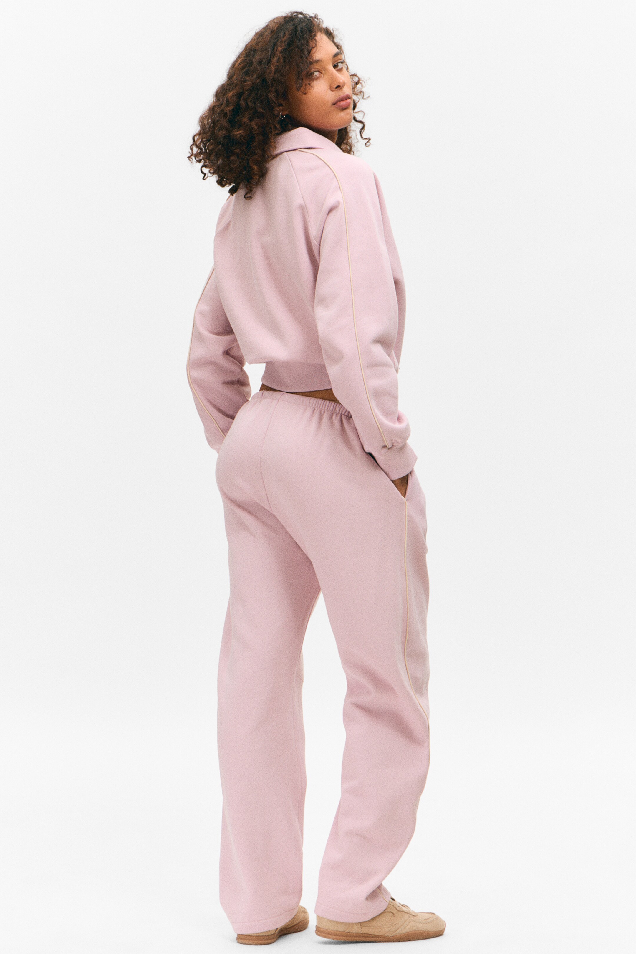 Agrandir l'image: Le survêtement rose est présenté d'une vue arrière trois-quarts. Le haut court présente un col et un passepoil de couleur claire s'étendant le long des manches, tandis que le pantalon à jambes larges assorti affiche une taille élastiquée et un passepoil de couleur claire courant le long des coutures latérales.