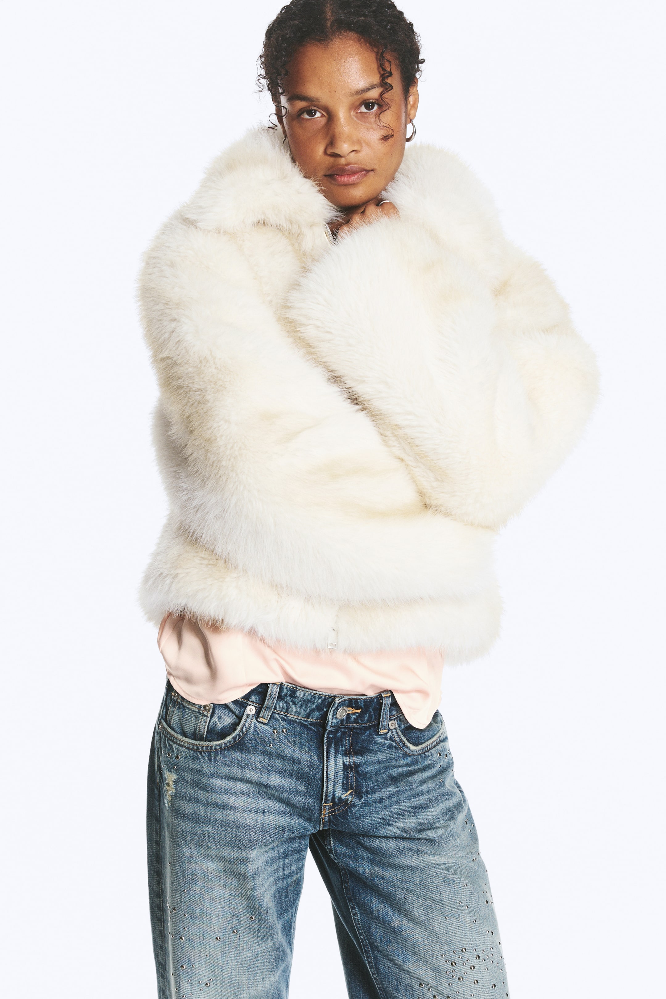 Größeres Bild ansehen: Eine cremeweiße Kunstpelzjacke mit breitem Kragen wird vom Model getragen, das die Arme eng vor der Brust verschränkt. Die kurze Jacke ist über einem hellrosa Oberteil getragen und mit blauen Jeans kombiniert.
