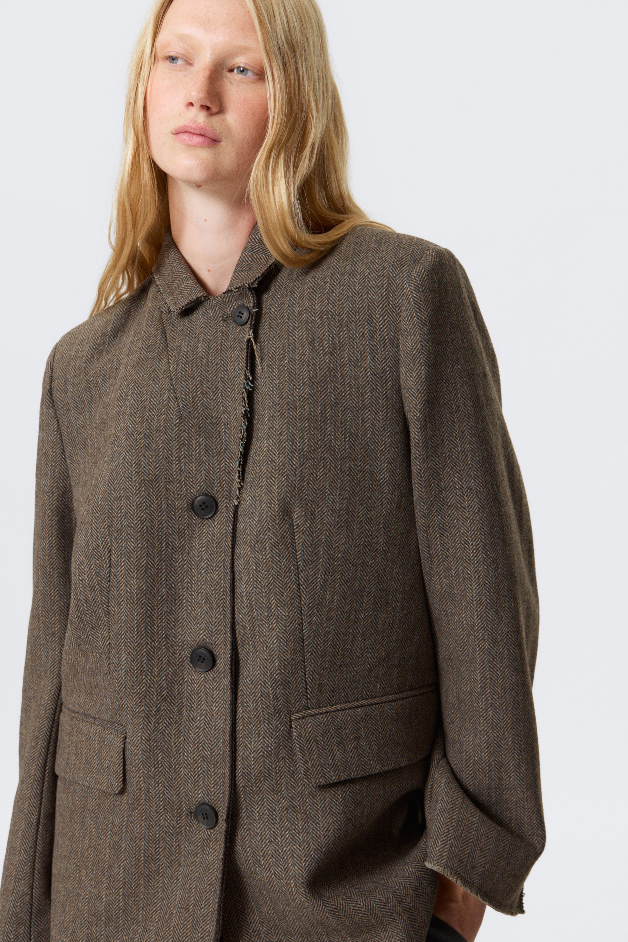 Agrandir l'image: Une veste en tweed marron à chevrons est présentée sous un angle légèrement incliné, avec un col pointu et une fermeture à trois boutons sur le devant. La veste présente deux poches à rabat sur le bas du devant, et des bords non finis et effilochés détaillent le col, la patte de boutonnage et les poignets.