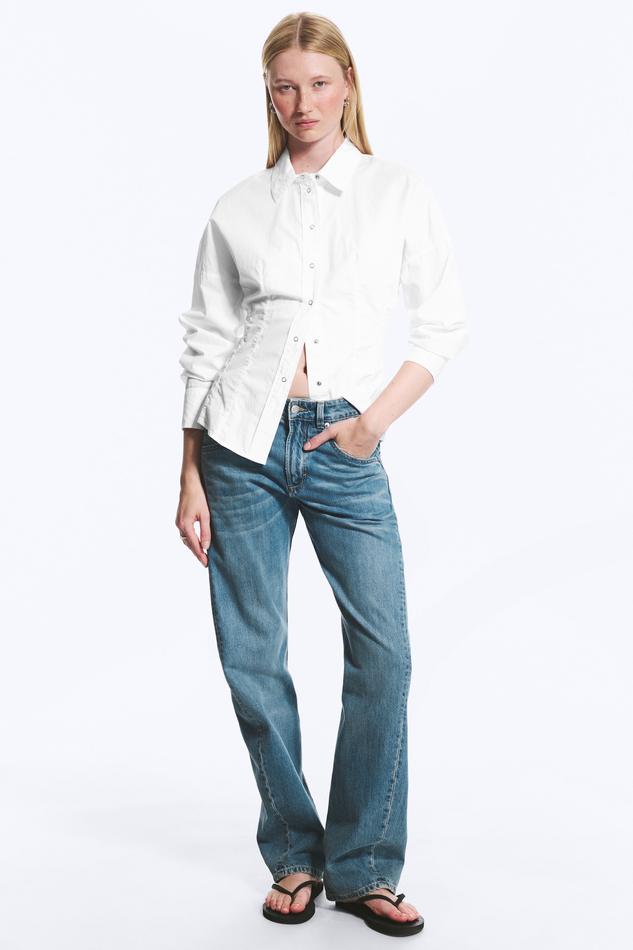 Ver imagen más grande: La camisa blanca de botones presenta un cuello en punta y mangas largas. Destaca por sus detalles prominentes de cintura fruncida y ajustada en ambos lados, con botones de presión que cierran la parte delantera, algunos de los cuales están desabrochados en la parte inferior.