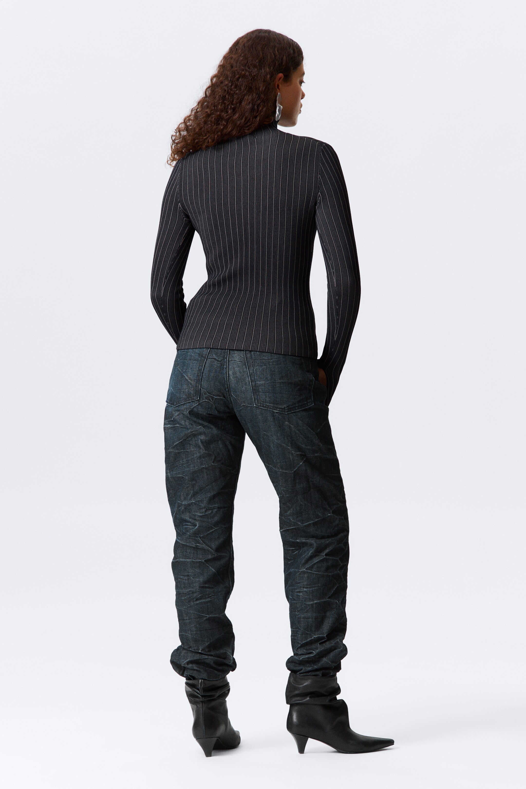 Größeres Bild ansehen: Ein schwarzes, enganliegendes, langärmliges Oberteil mit feinen weißen vertikalen Nadelstreifen wird von hinten gezeigt, kombiniert mit dunklen Jeans, die starke Knitterfalten aufweisen und eine lange Länge haben, die sich über schwarzen spitzen Stiefeln staut.