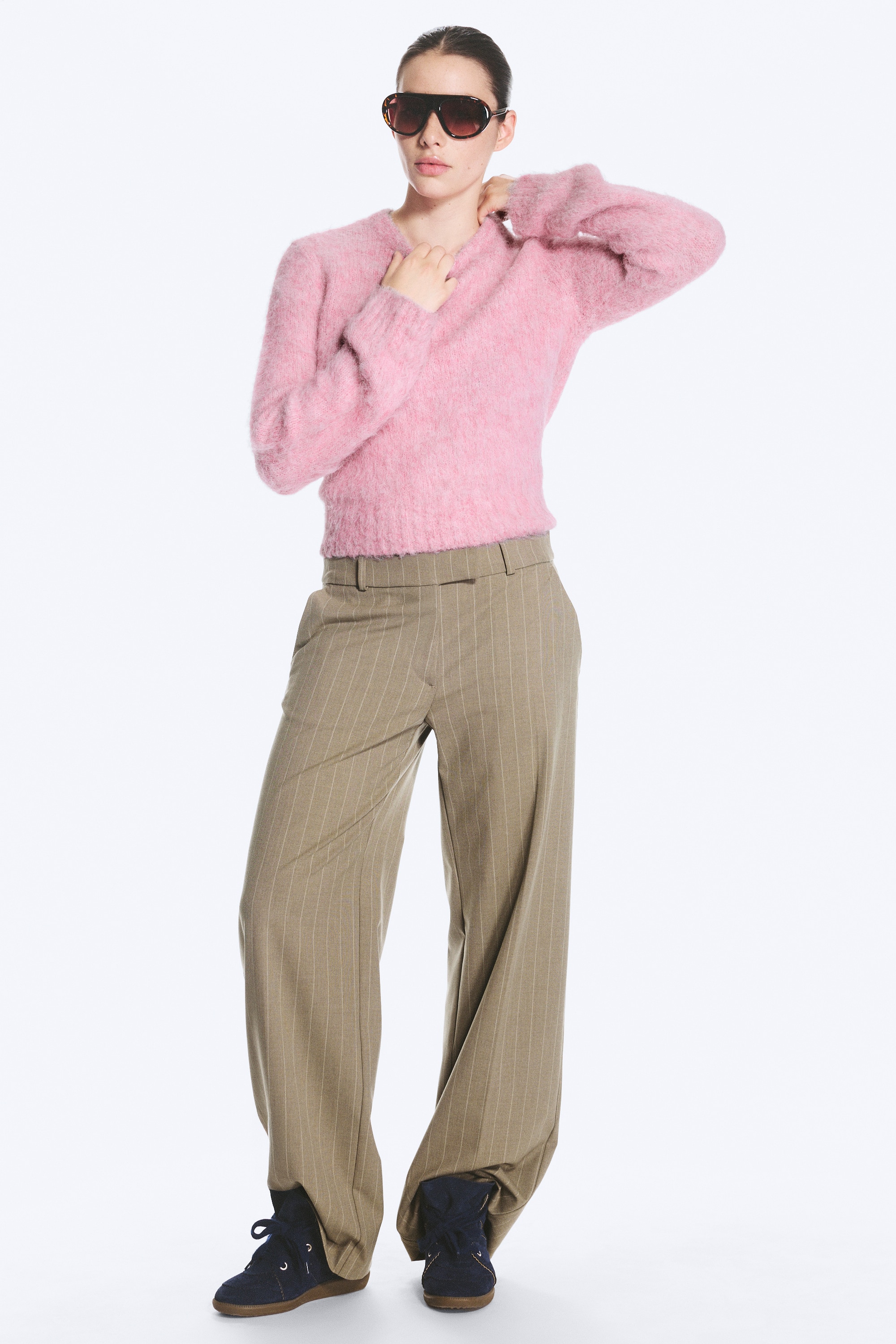 Een paar beige laagzittende broek met krijtstrepen en een rechte pijp, gemaakt van gestructureerde stof, gezien vanaf de voorkant. Ze zijn voorzien van schuine zijzakken, geperste vouwen die over de voorkant lopen, en een nette tailleband met een haak-en-oog sluiting.
