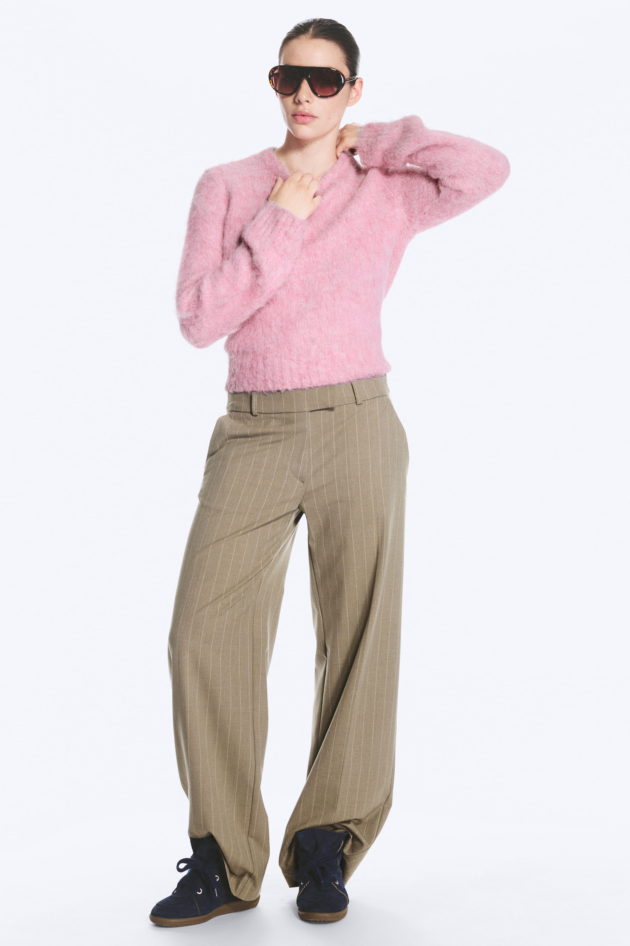 Größeres Bild ansehen: Der Model trägt eine sandfarbene Anzughose mit feinen Nadelstreifen, kombiniert mit einem flauschigen rosa Langarmpullover und dunkel gefärbten Schnürschuhen. Die Hose mit niedriger Leibhöhe verfügt über eine entspannte, gerade Passform, schräge Seitentaschen und einen sauberen Bund mit Gürtelschlaufen.