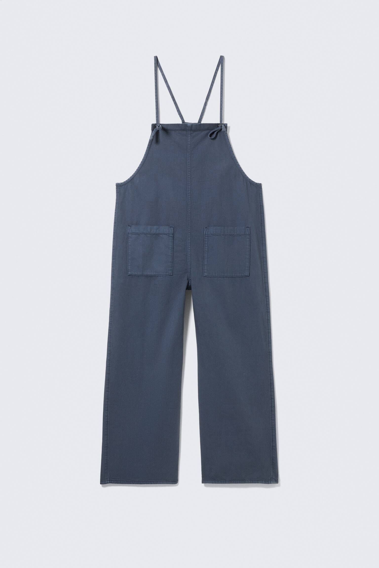 Dark blue dungarees are shown from the back, featuring two thin shoulder straps that cross and tie at the upper back. Two large, rectangular patch pockets are visible on the seat, and wide-leg trousers extend downwards.