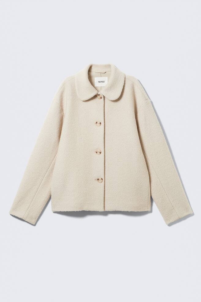 A cream-colored jacket is being shown from the front, displaying a textured bouclé fabric. It features a rounded Peter Pan collar and three light brown buttons closing the front. - 1