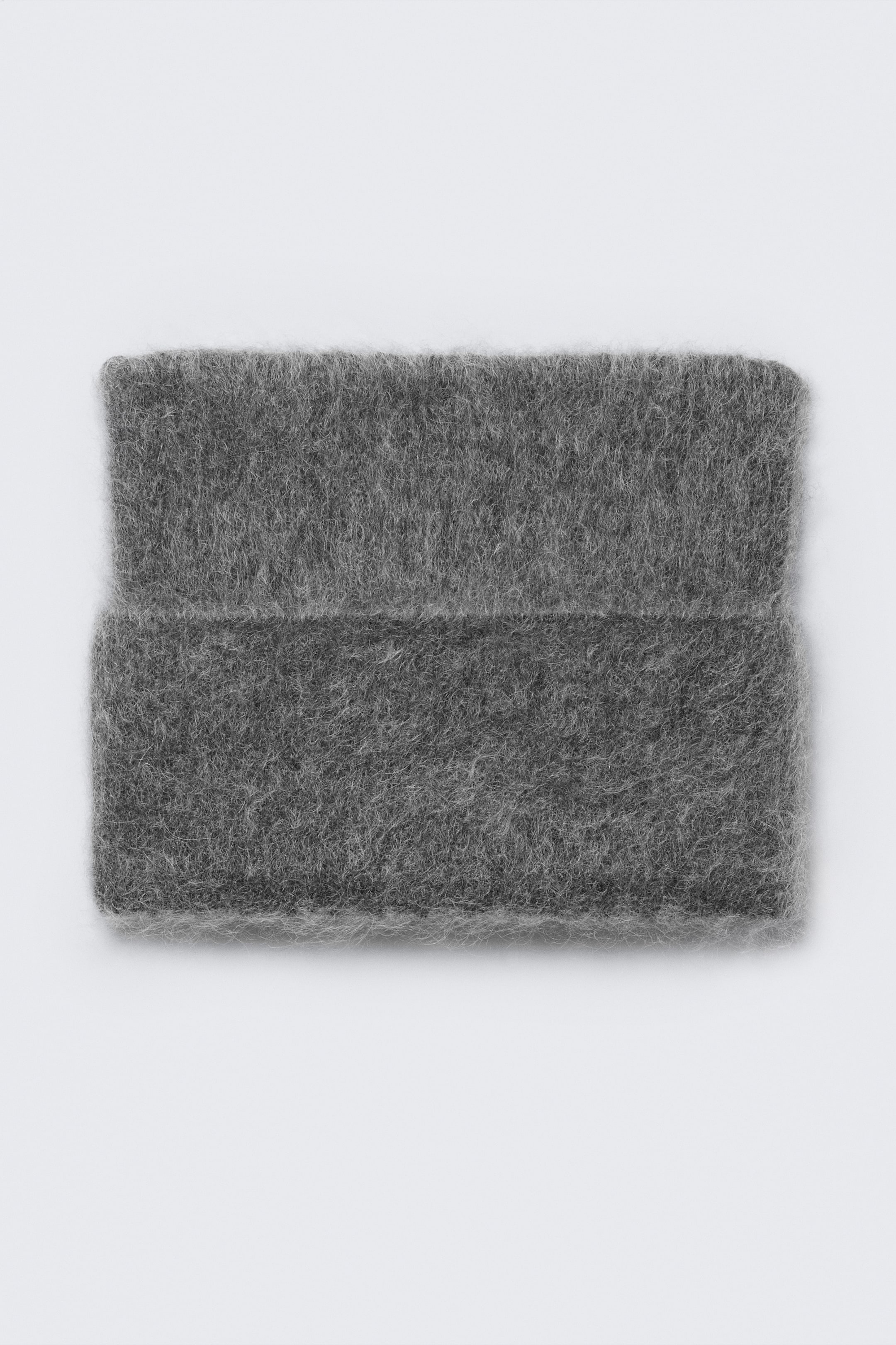 Visa större bild: En grå, luddig stickad mössa presenteras framifrån, vikt platt, med en uttalad borstad textur och en synlig horisontell veck.