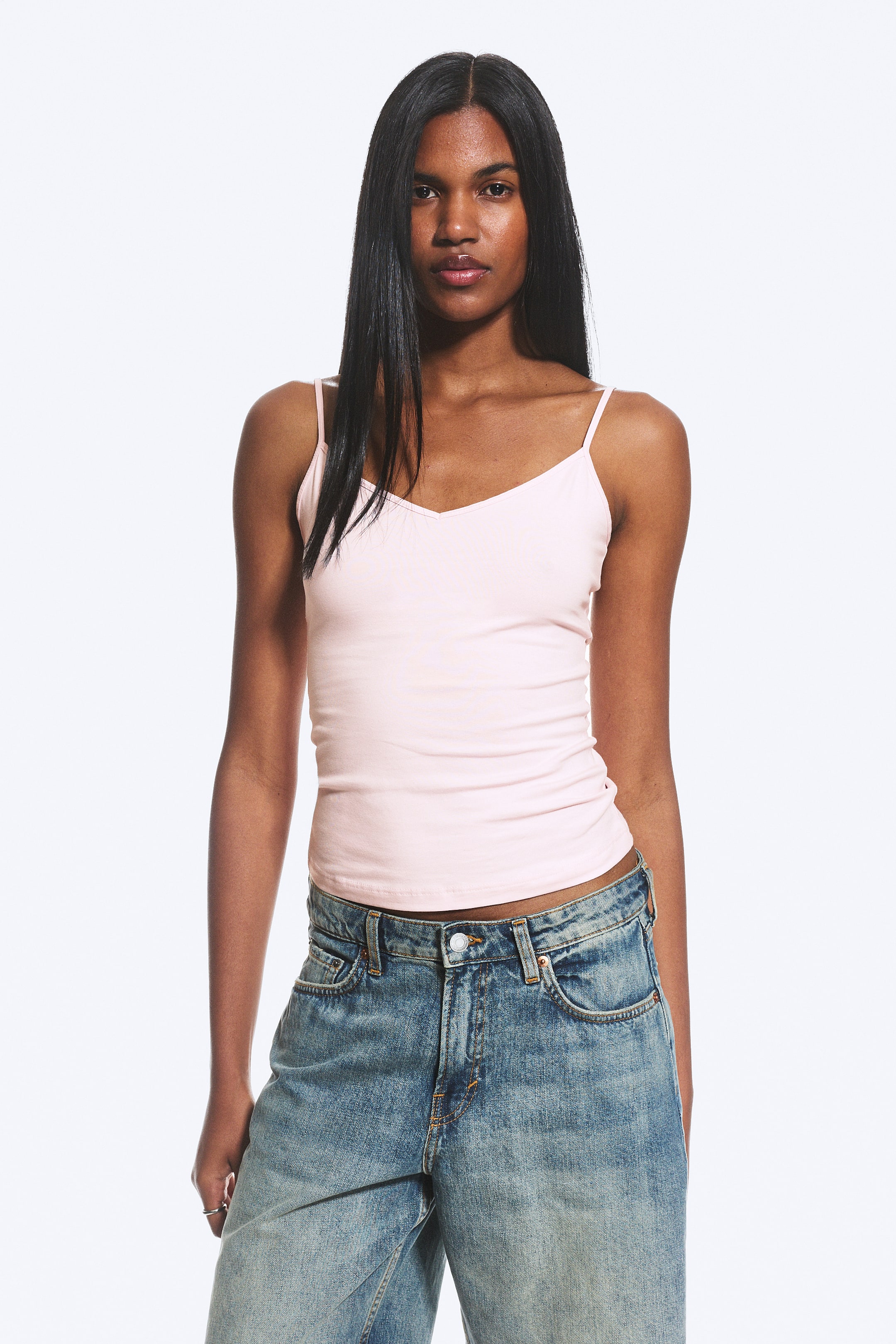 Ver imagem maior: Uma camisola de alças em rosa claro é apresentada de frente, com um decote em V, finas alças spaghetti e uma textura de malha subtilmente canelada, contribuindo para a sua silhueta ajustada.