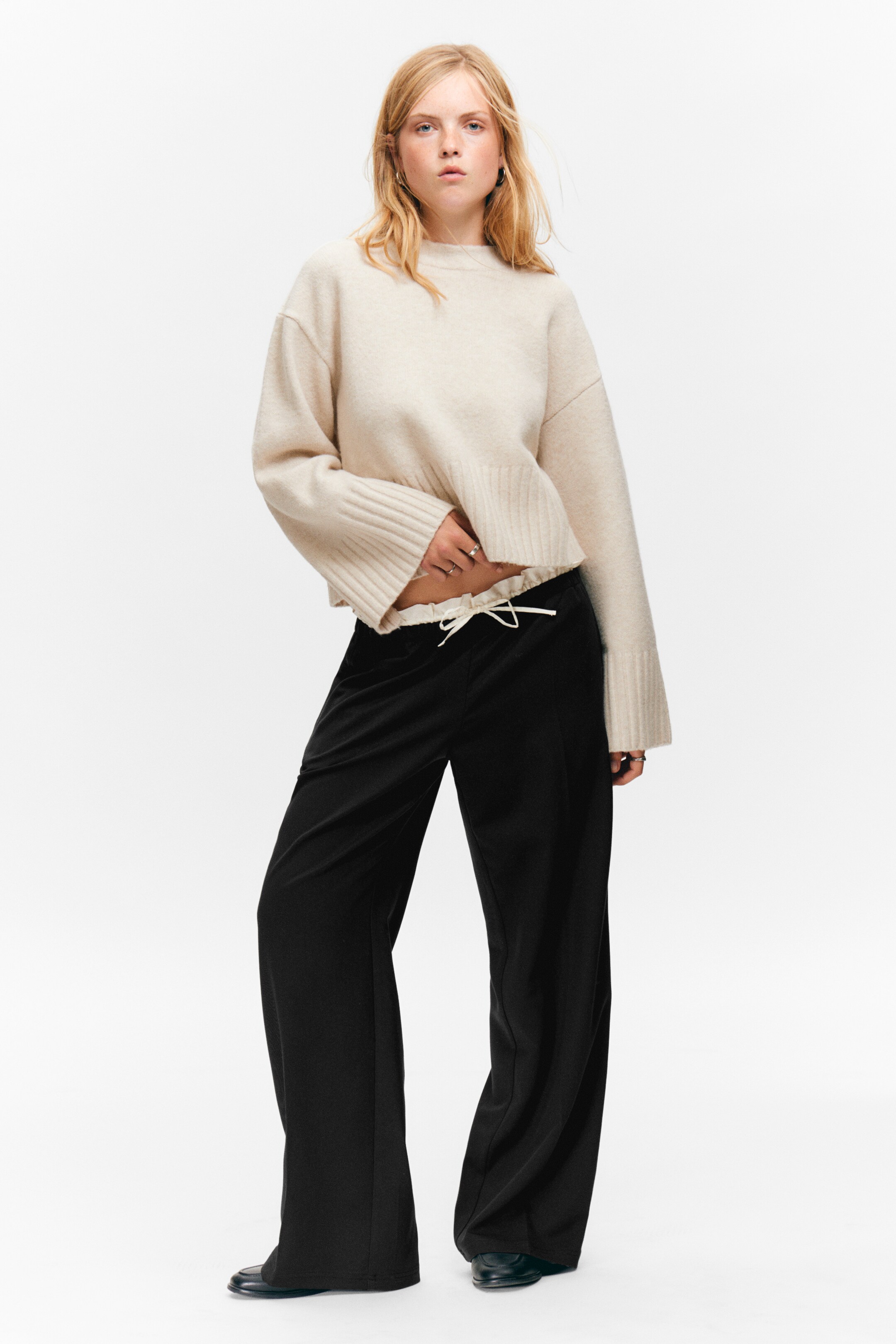 Visualizza immagine più grande: Un maglione beige chiaro, a maniche lunghe e con scollo rotondo, appare con una vestibilità rilassata, polsini a coste larghe e un orlo a coste e arricciato che rivela il cordoncino bianco dei pantaloni neri a gamba larga. I pantaloni presentano una vestibilità rilassata e a lunghezza intera.