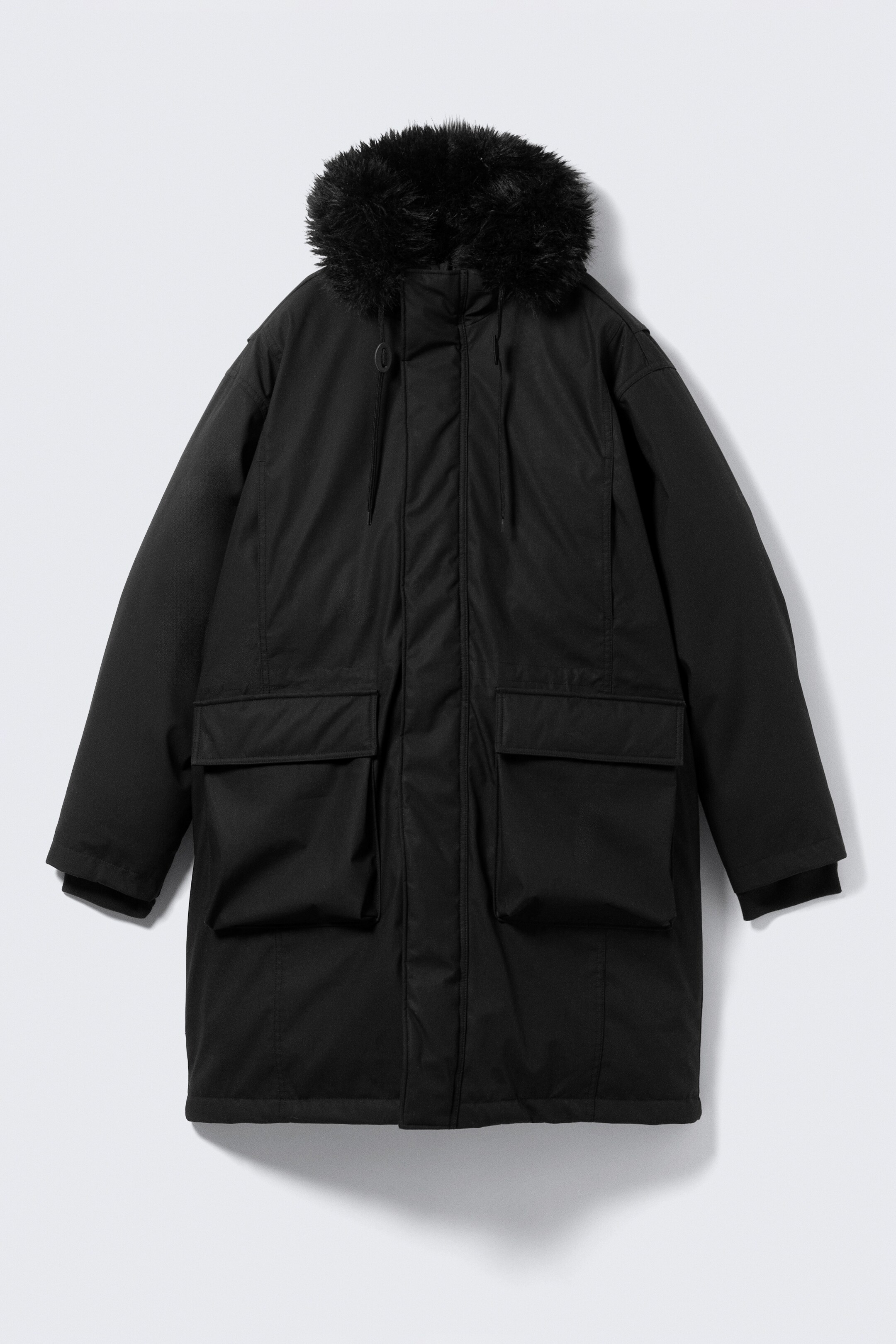 Grotere afbeelding bekijken: Een zwarte oversized parka die tot over de knieën reikt, wordt VOOR gesteld, met een gewatteerde constructie, een hoge kraag en een kap afgezet met zwart nepbont en een verstelbaar trekkoord. De voorkant toont een ritssluiting die verborgen is door een drukknoopsluiting, twee grote klepzakken, en stormmanchetten zijn zichtbaar aan de mouwen.