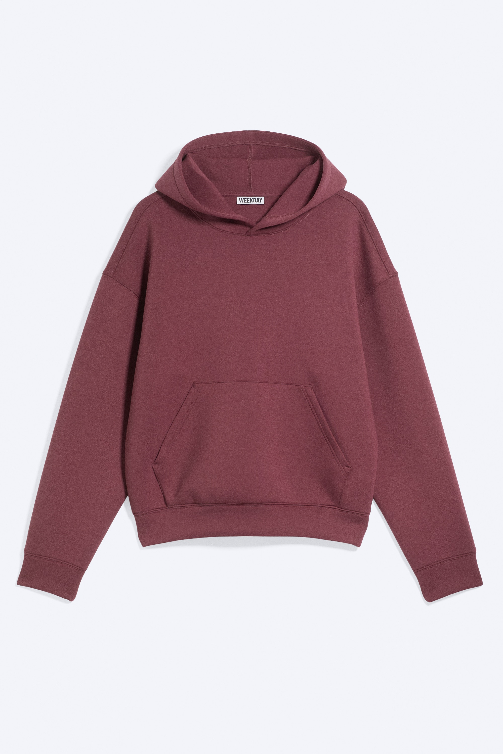A burgundy loose-fit hoodie in a structured scuba fabric is being worn by the model, who has both hands in the front kangaroo pocket. The hoodie features dropped shoulders and is layered over a white T-shirt.