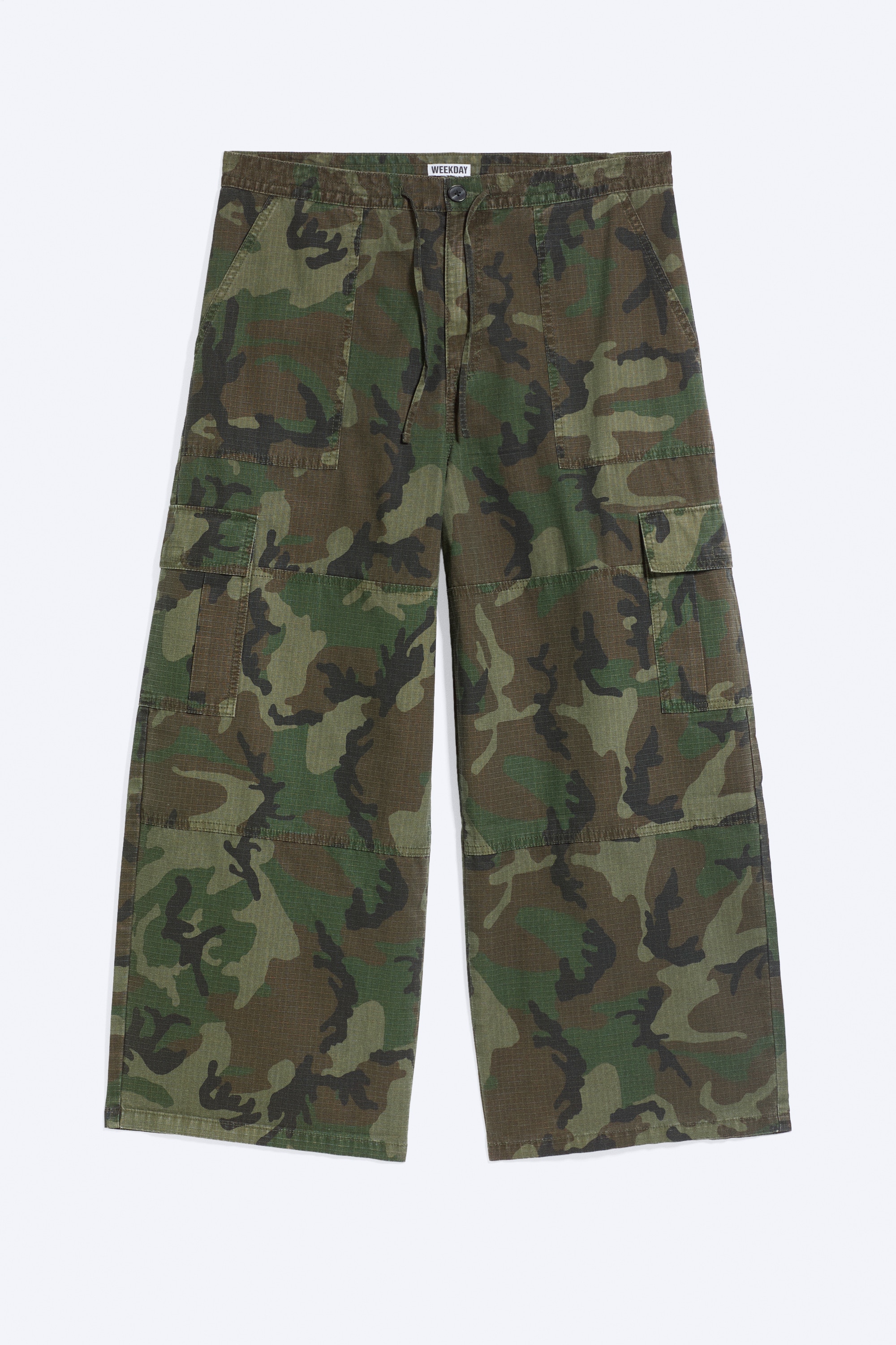Vide cargo-bukser i ripstop med grÃ¸nt og brunt kamuflasjemÃ¸nster og lÃ¸s, vide ben bÃ¦res av modellen. Buksen er stylet med en lysegul T-skjorte, en mÃ¸rk crossbody-veske og hvite sneakers.