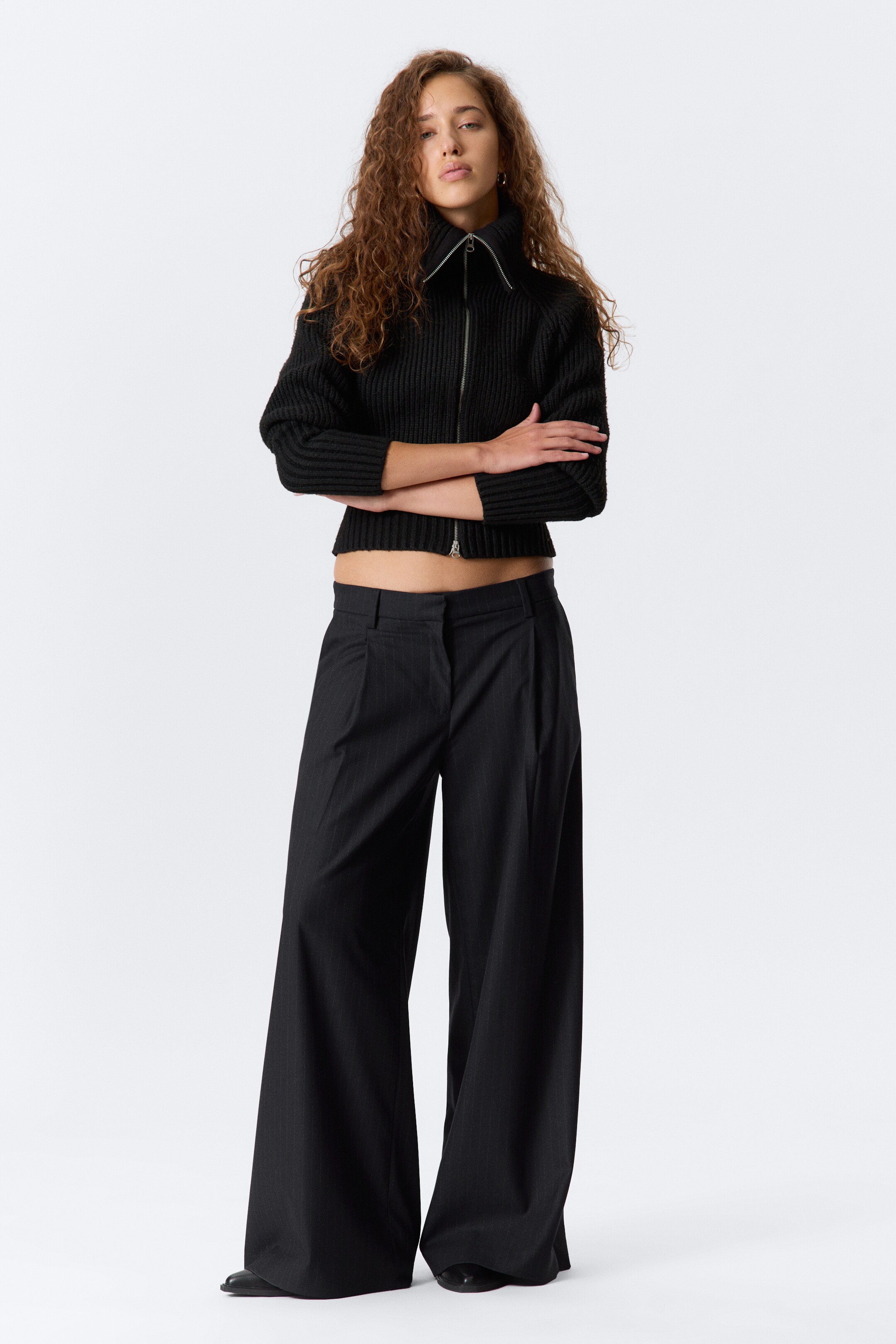 Grotere afbeelding bekijken: Een zwarte geribde cropped trui, met een volledige ritssluiting en een hoge kraag, wordt gecombineerd met zwarte wijd uitlopende broek met krijtstreep en plooien aan de voorkant, gepresenteerd met de voorkant naar voren.
