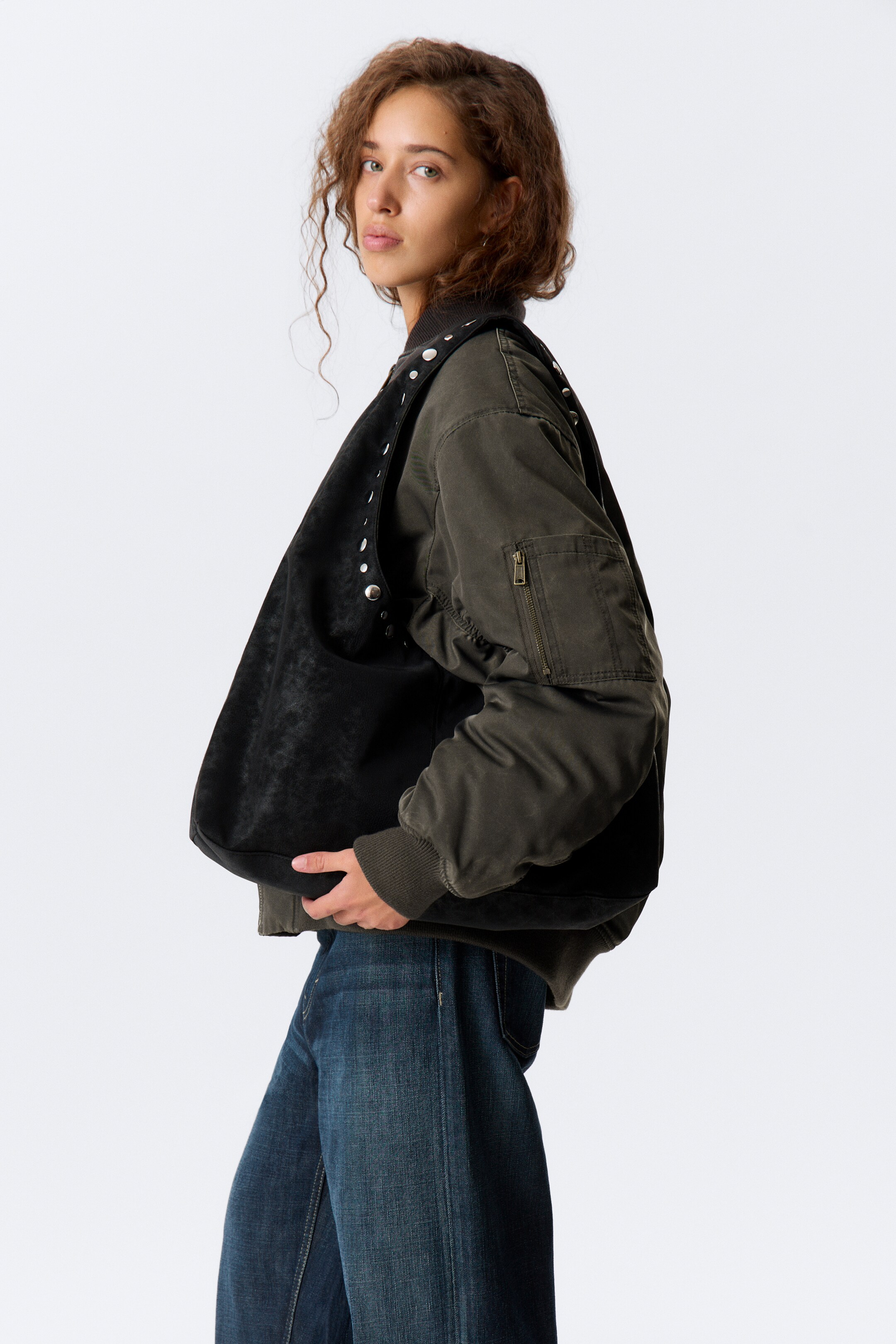 Agrandir l'image: Un sac à bandoulière en simili cuir noir, présentant une silhouette triangulaire douce et des détails en clous argentés le long de sa poignée supérieure arrondie, est porté par le mannequin sur son épaule droite. Le mannequin tient le bord inférieur avant du sac avec sa main droite, tout en portant un blouson aviateur olive.