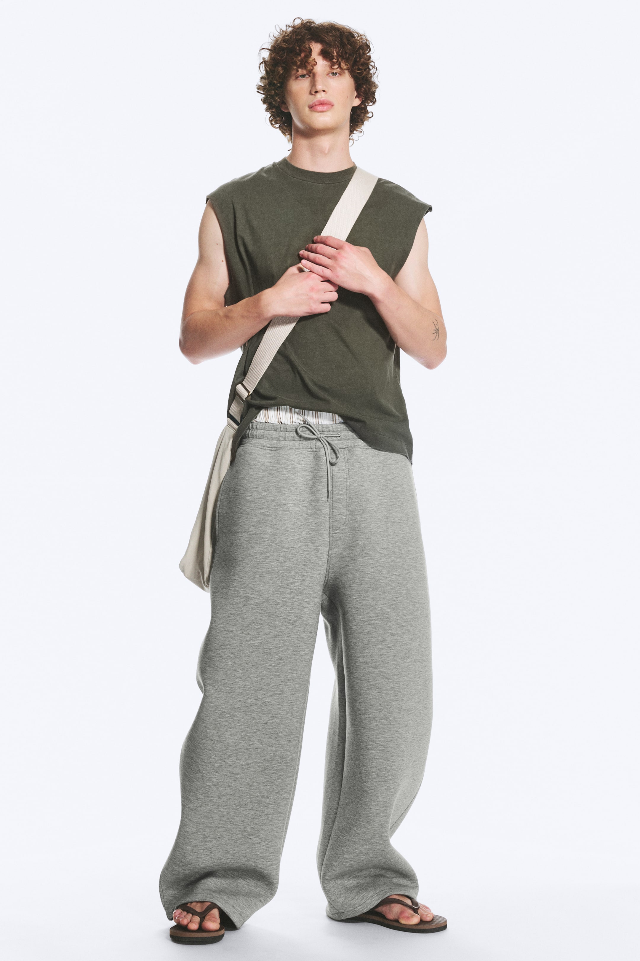 Größeres Bild ansehen: Der Model trägt die Cody Scuba Jogginghose in Melange-Grau. Sie zeichnet sich durch einen entspannten, weit geschnittenen Beinverlauf aus und verfügt über einen sichtbaren, elastischen Kordelzugbund, der gebunden ist. Die lange Hose fällt leicht über braune Flip-Flops, und ein gestreifter Unterwäschebund ist knapp oberhalb der Jogginghose sichtbar.