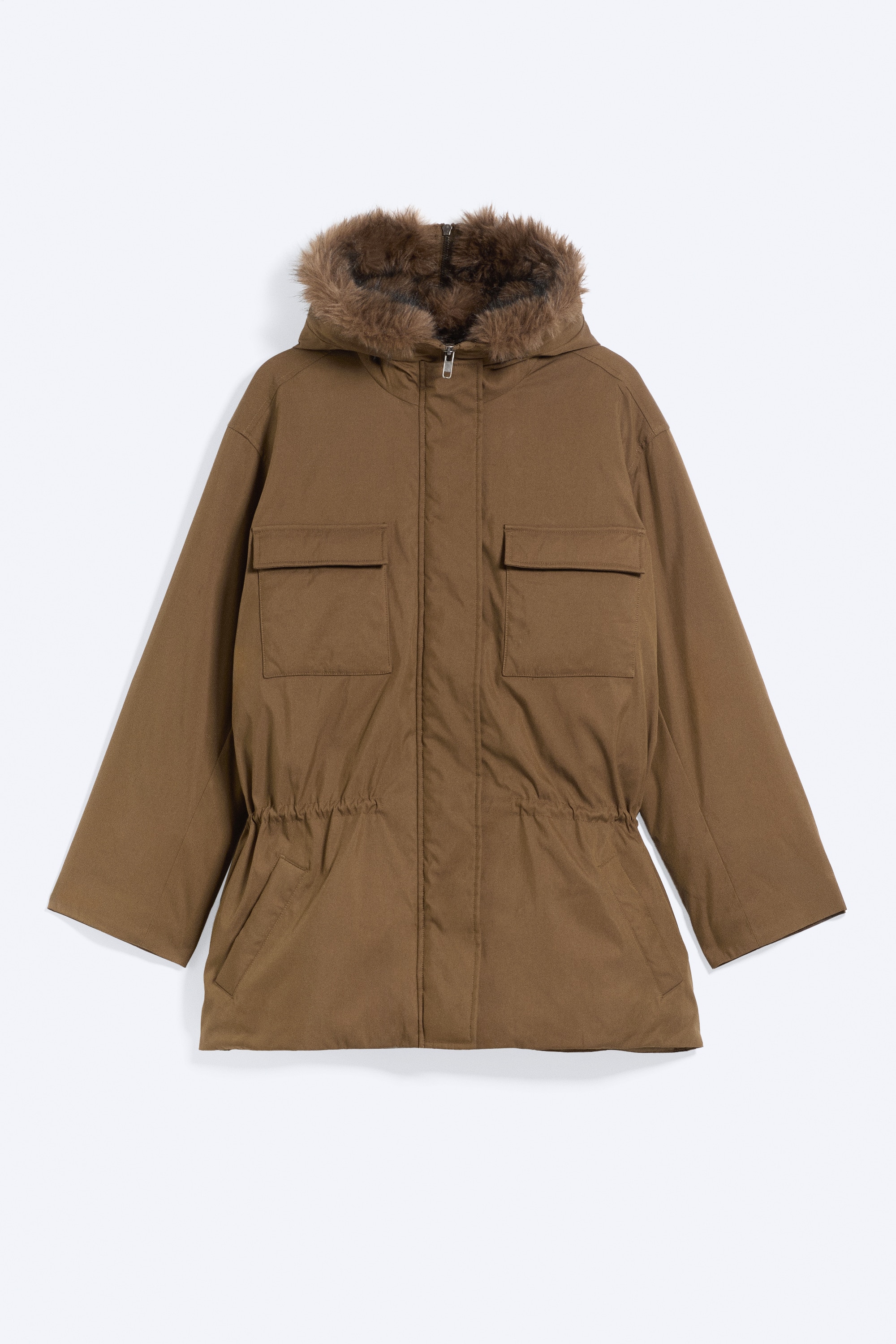 A brown cotton canvas parka featuring a faux-fur lined hood split open into a collar is worn by the model over a cream top and ribbed knit trousers. The jacket is styled open to show a cinched drawstring waist and a chest flap pocket, with the model placing one hand in a side pocket.