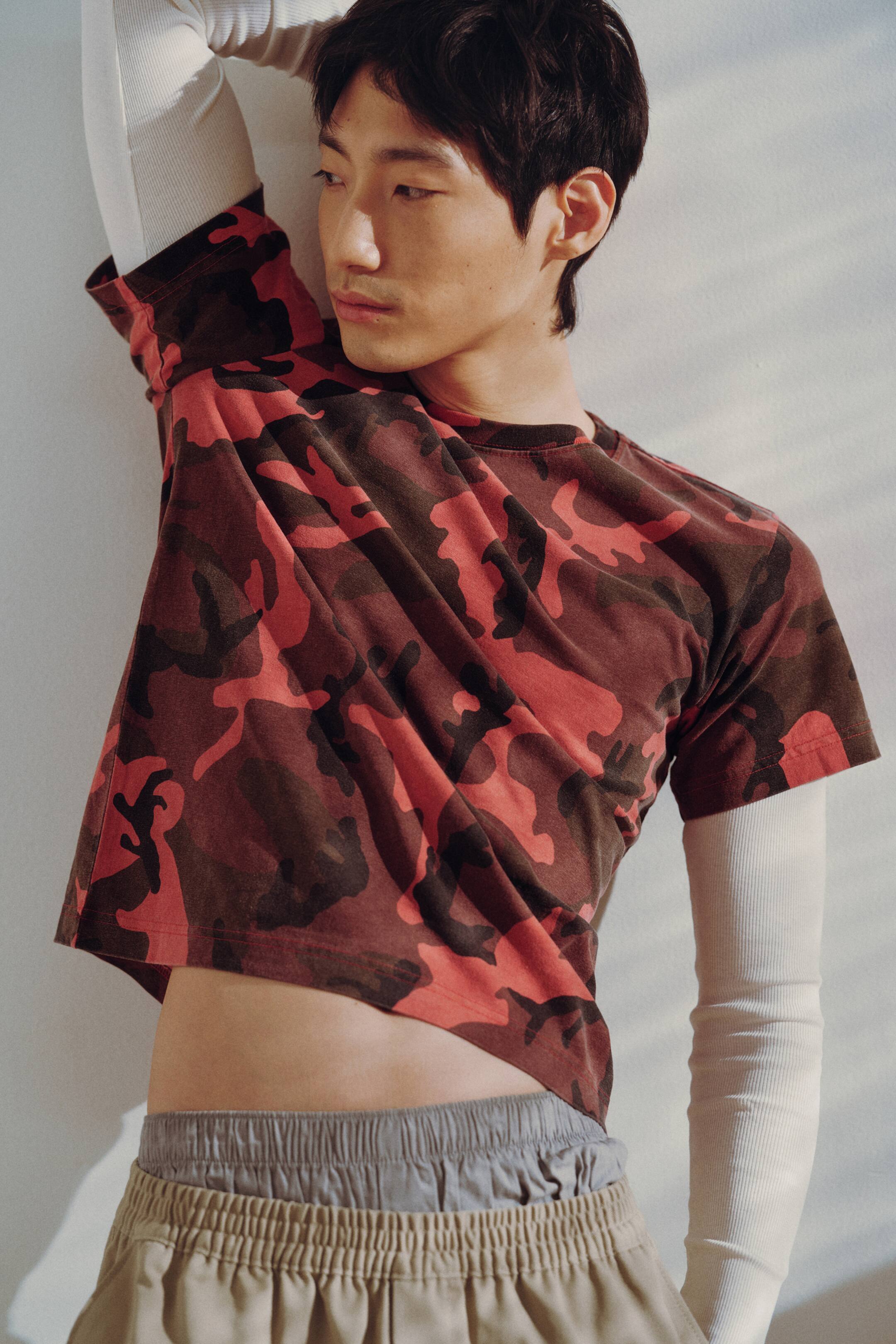 Agrandir l'image: Un t-shirt court est porté, arborant un motif camouflage rouge et brun foncé, un col rond et des manches courtes. Il est superposé à un maillot à manches longues côtelé de couleur claire.