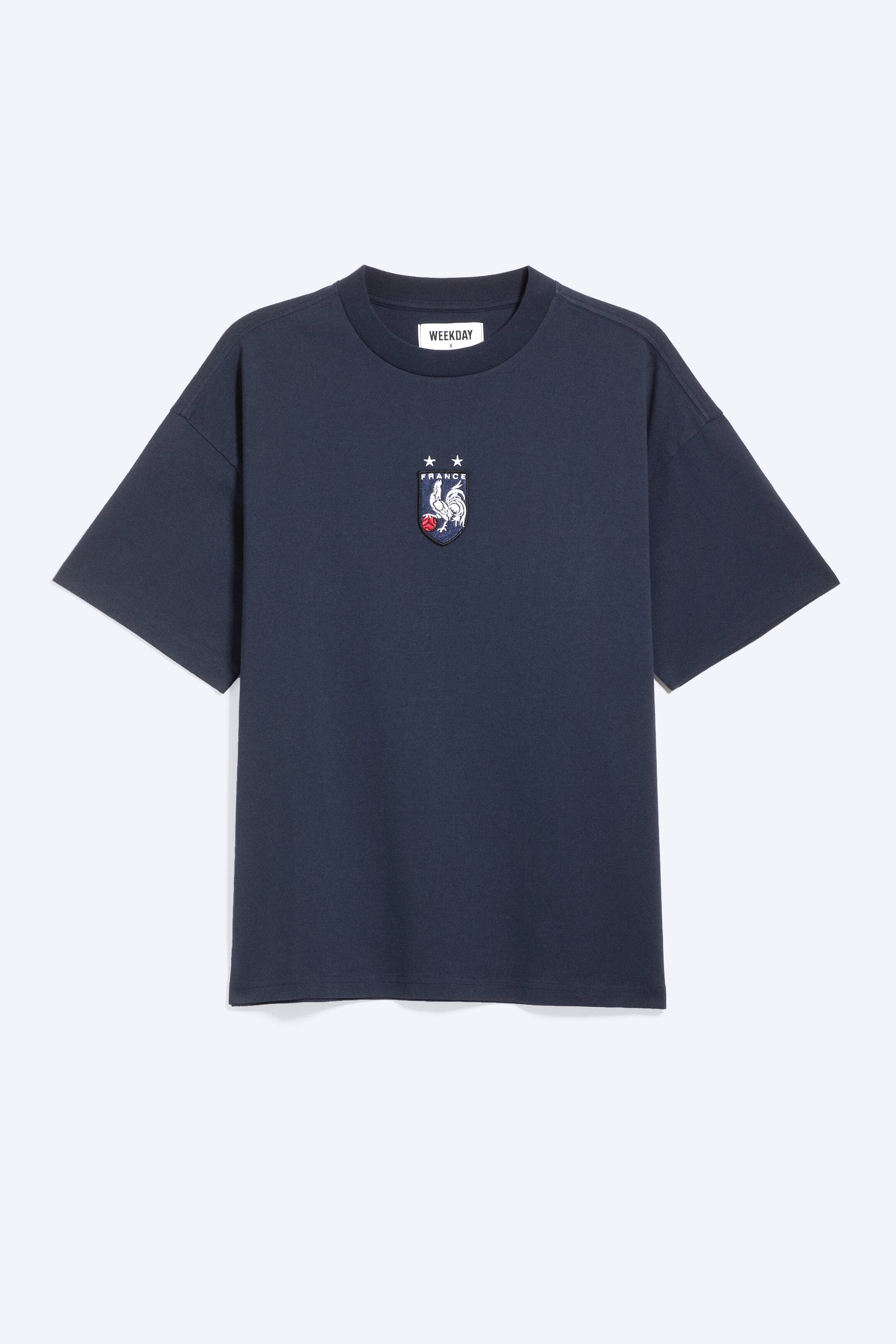Ver imagen más grande: Una camiseta azul marino presenta su vista frontal, con un cuello redondo acanalado y mangas cortas y relajadas. Un parche bordado en forma de escudo adorna el pecho izquierdo, mostrando un gallo blanco, una pelota roja y la palabra "FRANCE" con dos estrellas encima.