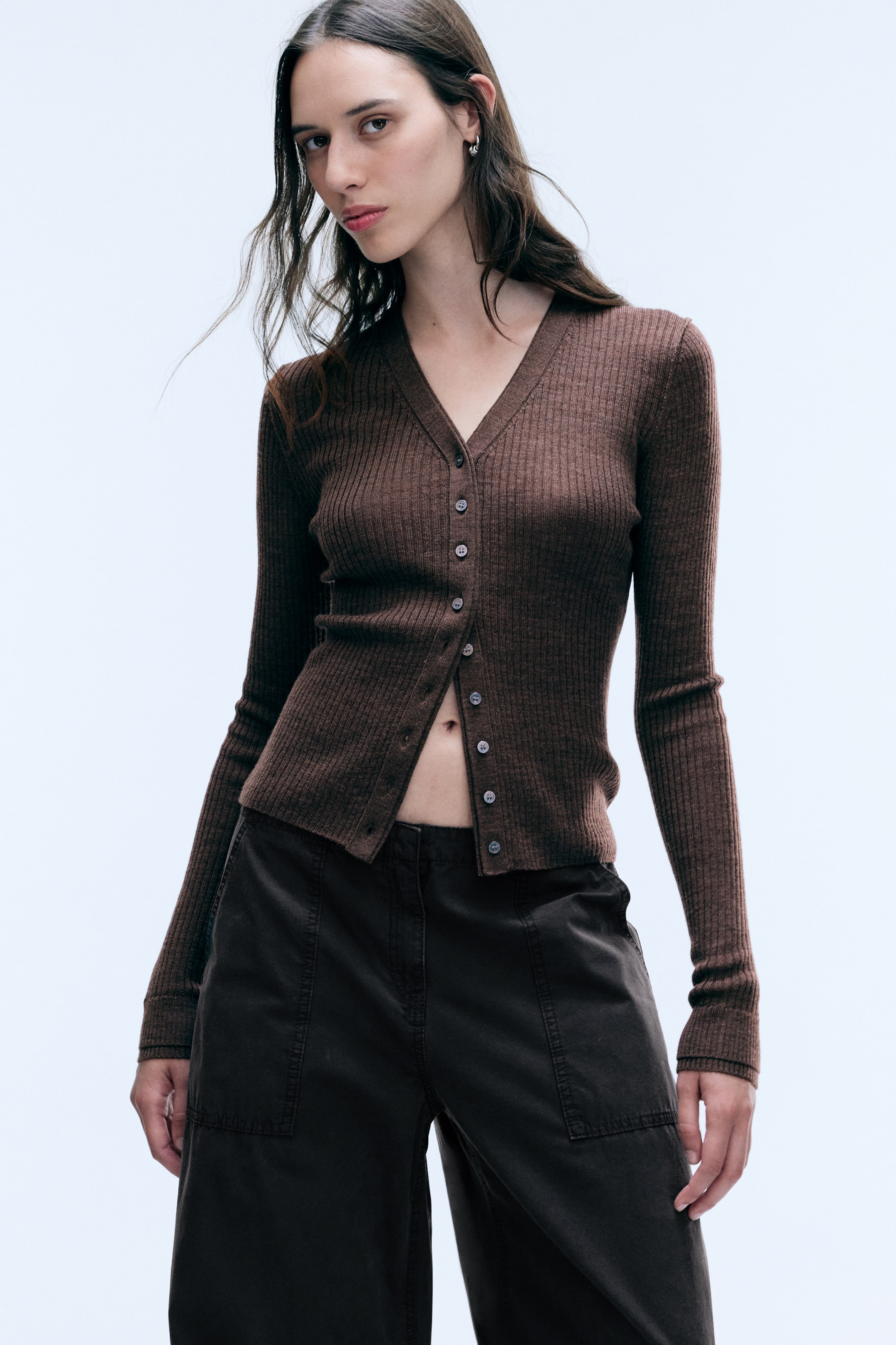 Vezi imaginea mai mare: Un cardigan din lână merinos, maro, tricotat cu model cu nervuri, cu decolteu în V și mâneci lungi, este purtat de model. Cardiganul are închidere frontală cu nasturi, parțial descheiată în partea de jos, și este asortat cu pantaloni închiși la culoare, cu croială largă.