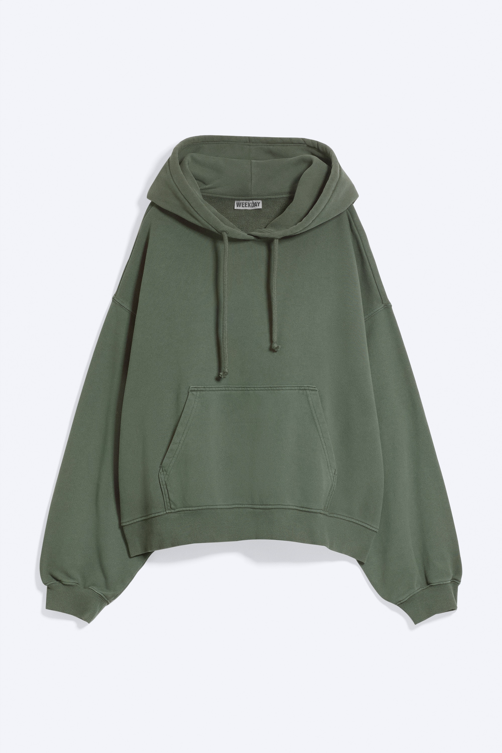 Una felpa con cappuccio verde oversize, con cappuccio con coulisse, spalle scese e orlo a costine, indossata dalla modella. La modella è in piedi con le mani infilate nella tasca a marsupio frontale.
