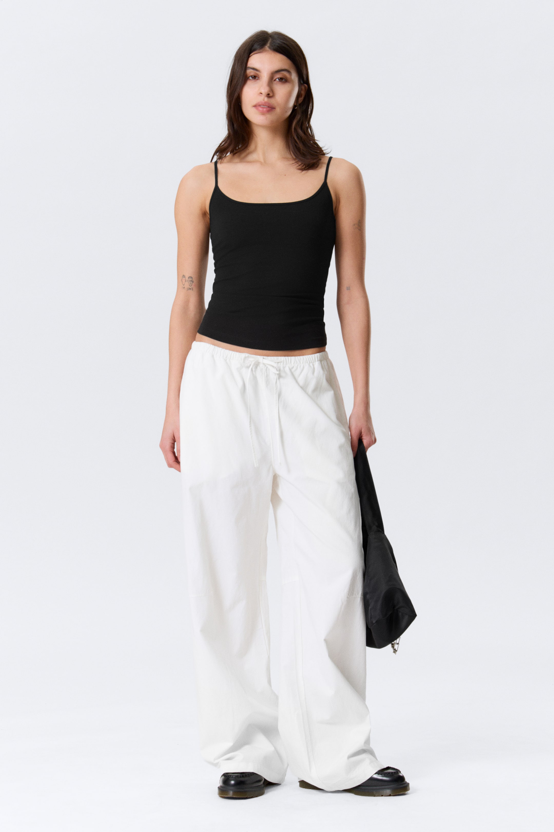 Grösseres Bild ansehen: Ein schwarzes, geripptes Camisole mit dünnen Spaghettiträgern und einem Rundhalsausschnitt wird präsentiert, kombiniert mit weissen, weit geschnittenen Hosen mit elastischem Kordelzugbund.