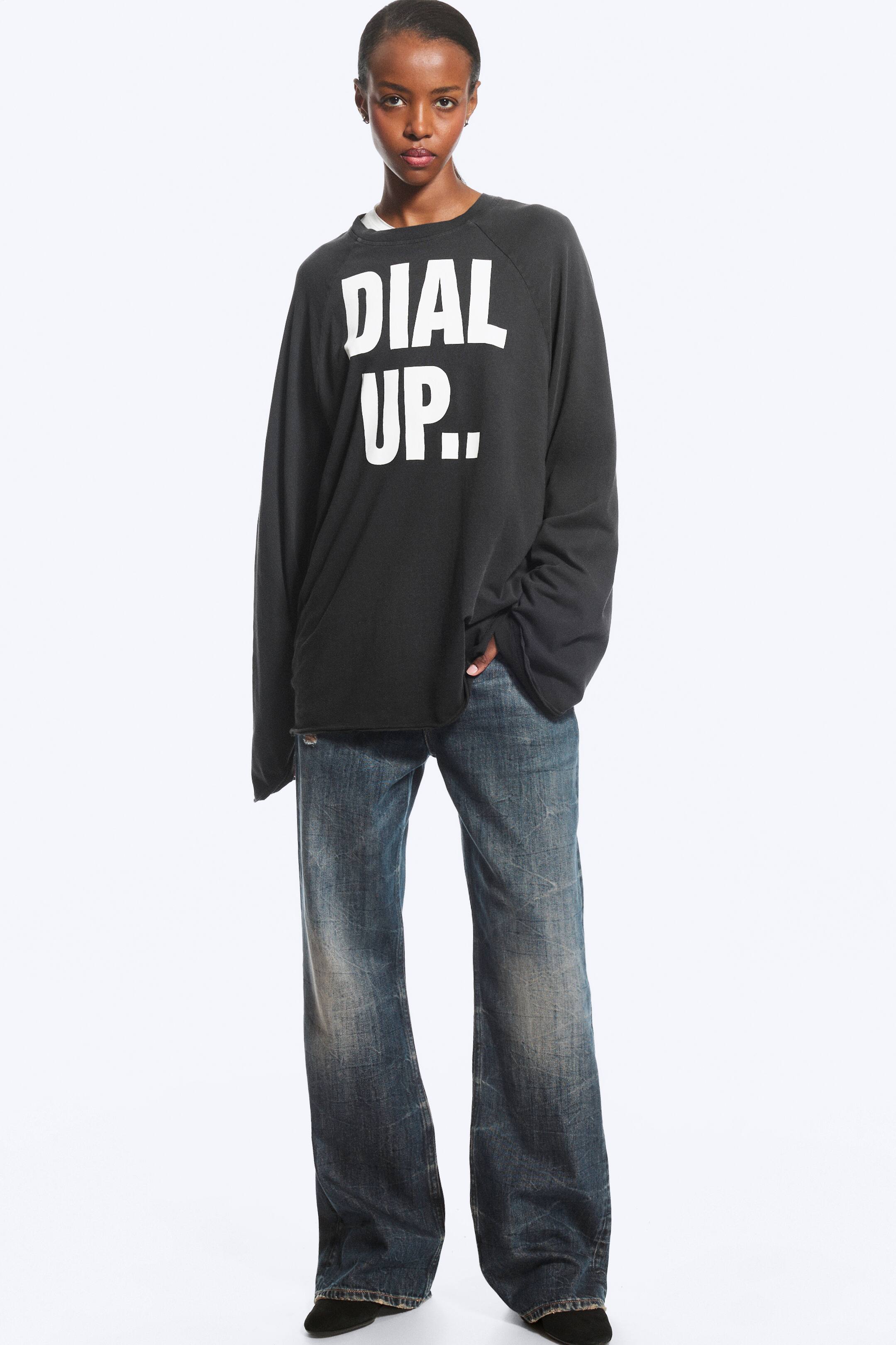 Agrandir l'image: Un sweat-shirt noir oversize à manches longues, orné du texte blanc en lettres capitales « DIAL UP.. » sur le devant, est porté par le modèle. Le sweat-shirt est associé à un jean bleu délavé à jambes larges.