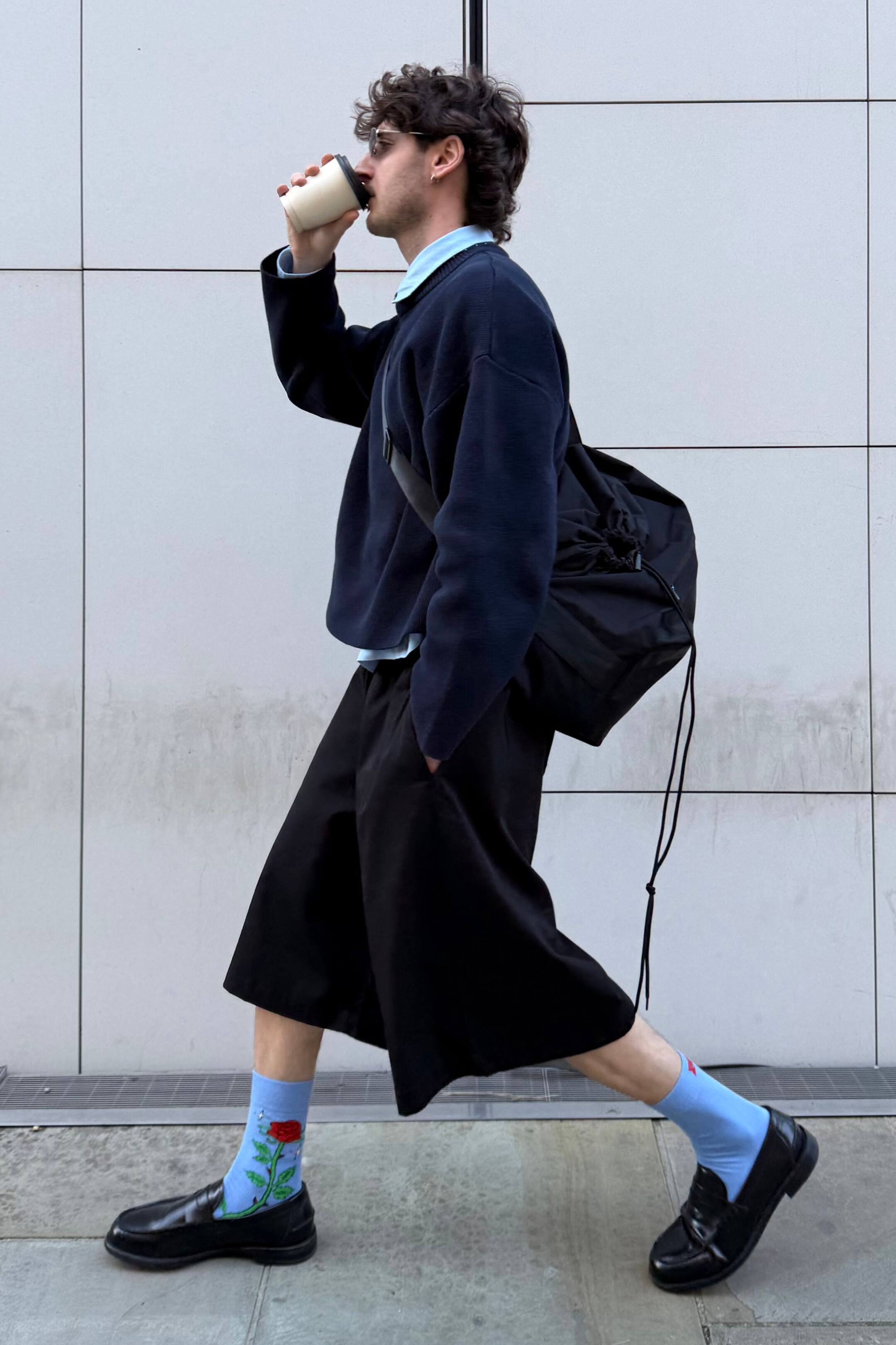 Agrandir l'image: Une personne porte un pull oversize bleu marine superposé sur une chemise à col bleu clair, un short noir à jambes larges et des mocassins noirs, le tout vu de profil en marchant. Des chaussettes bleu clair avec un motif de rose rouge sont visibles au-dessus des mocassins, et un sac noir à bandoulière avec cordon repose sur son dos.