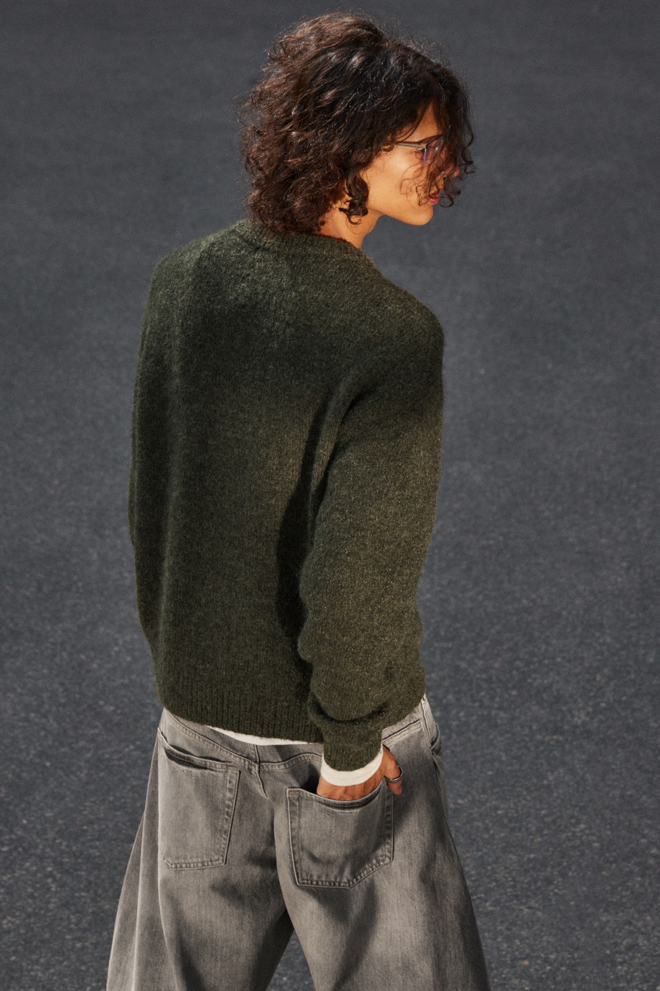 Größeres Bild anzeigen: Ein dunkelgrüner, flauschiger Strickpullover mit Rundhalsausschnitt, entspannter Passform und geripptem Saum und Bündchen wird getragen, von hinten und von der rechten Seite gesehen. Er ist über einem weißen Unterhemd geschichtet und mit ausgewaschenen grauen, weit geschnittenen Jeans mit sichtbaren Gesäßtaschen gestylt.