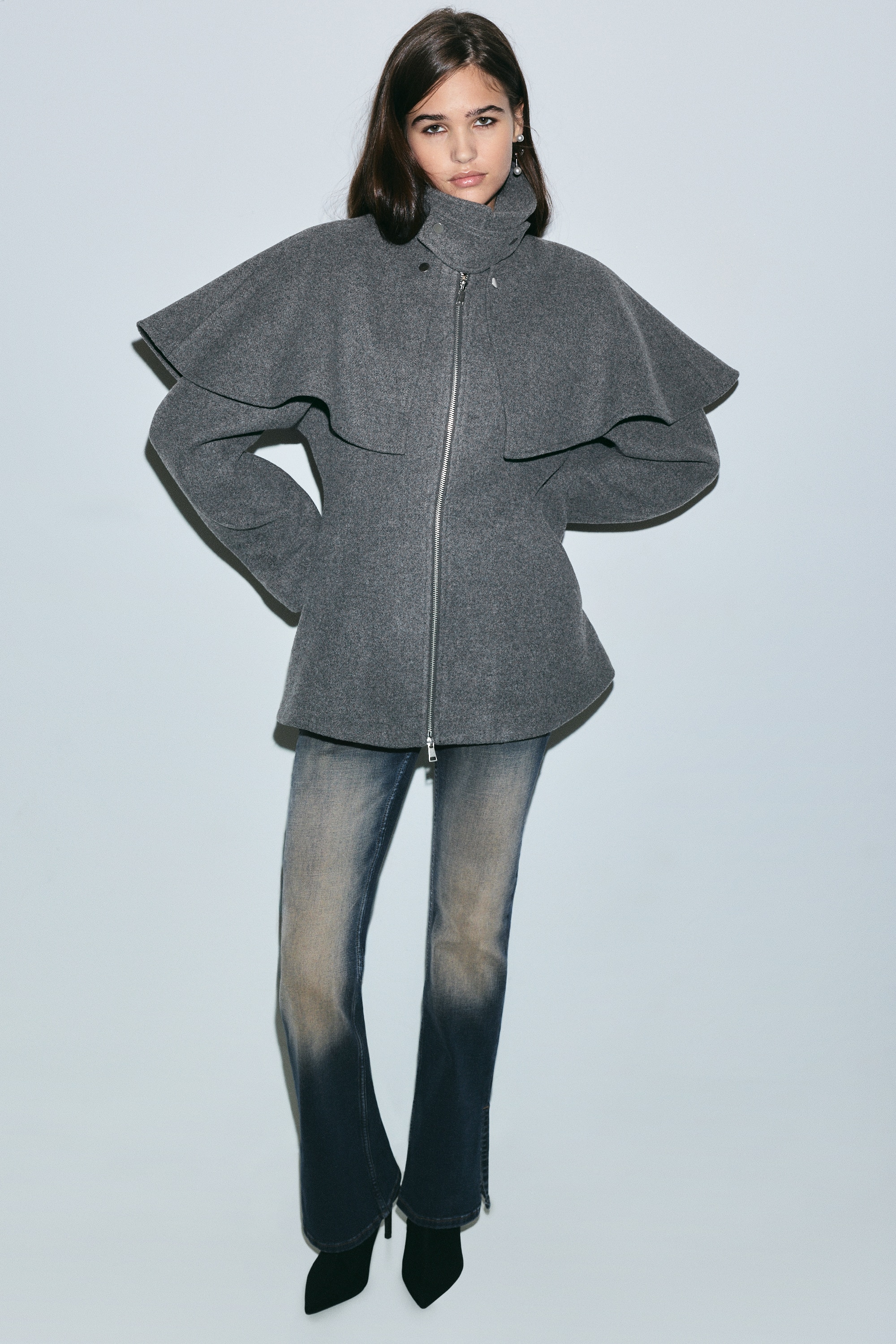 Se muestra una chaqueta de mezcla de lana gris desde el frente, con cuello alto tipo chimenea con trabilla abotonada en la garganta y una capa superpuesta a juego que cae sobre los hombros. La chaqueta está diseñada con cremallera frontal bidireccional, bolsillos ribeteados en ángulo y un bajo peplum ligeramente acampanado.