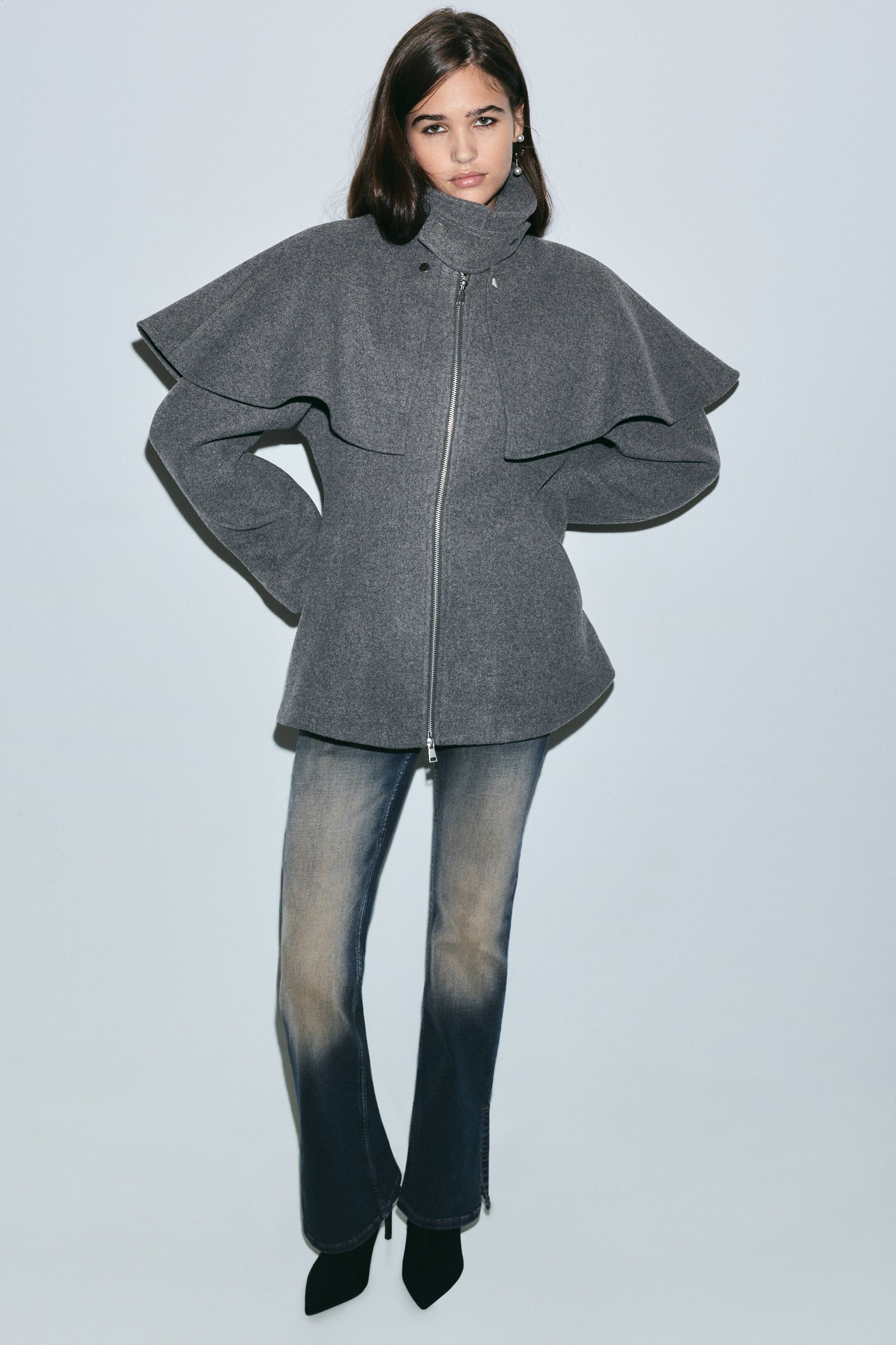 Ver imagen más grande: La modelo lleva una chaqueta gris con una amplia superposición tipo capa, cuello alto tipo chimenea y cierre de cremallera frontal. La chaqueta se combina con vaqueros azules desgastados y botas negras de punta afilada.