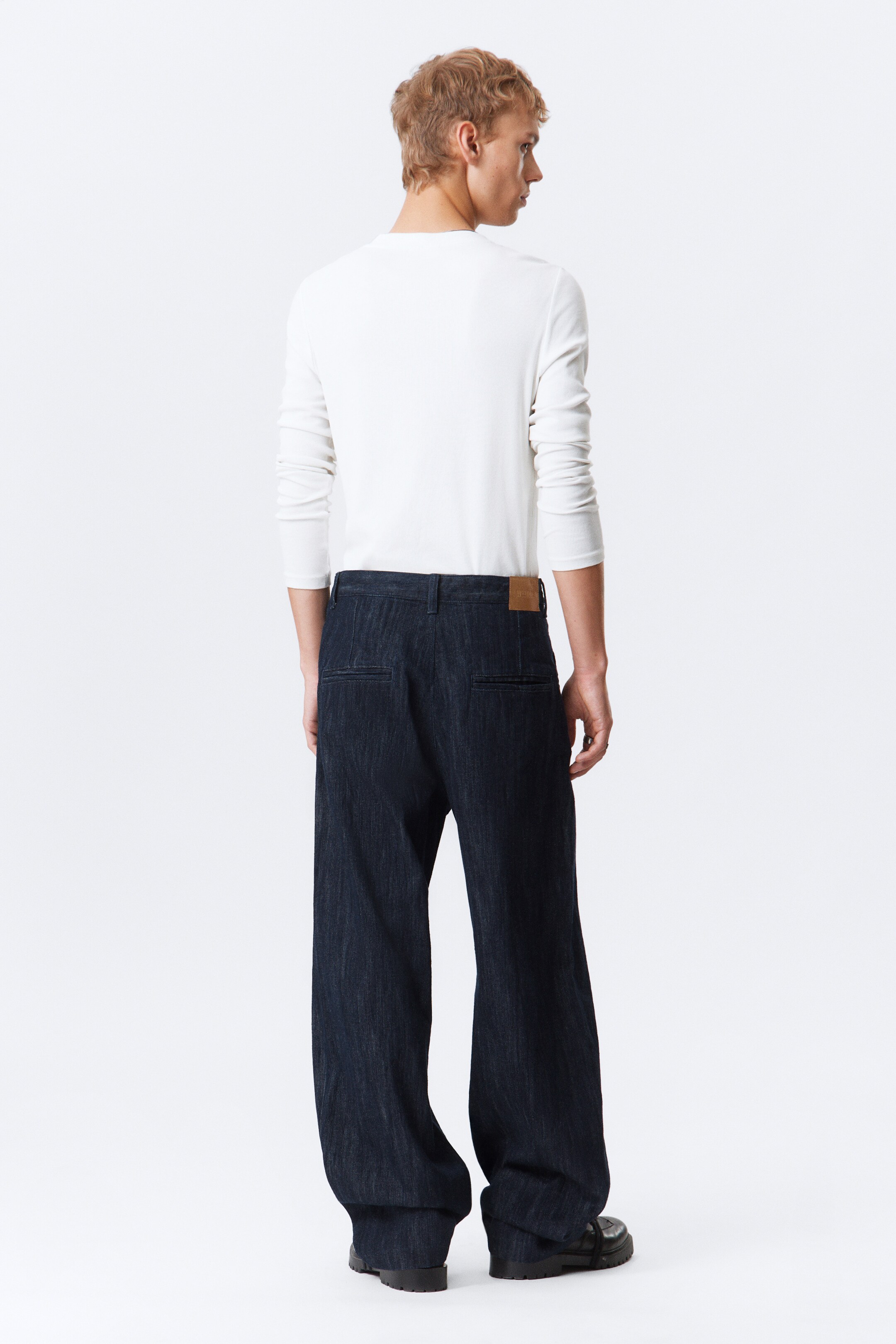 Agrandir l'image: Un pantalon large en denim foncé est orienté dos au spectateur, avec une coupe ample et une pièce marron clair apparaissant sur le côté droit de la ceinture arrière. Deux poches arrière sont visibles, et les ourlets larges tombent légèrement sur des chaussures foncées.