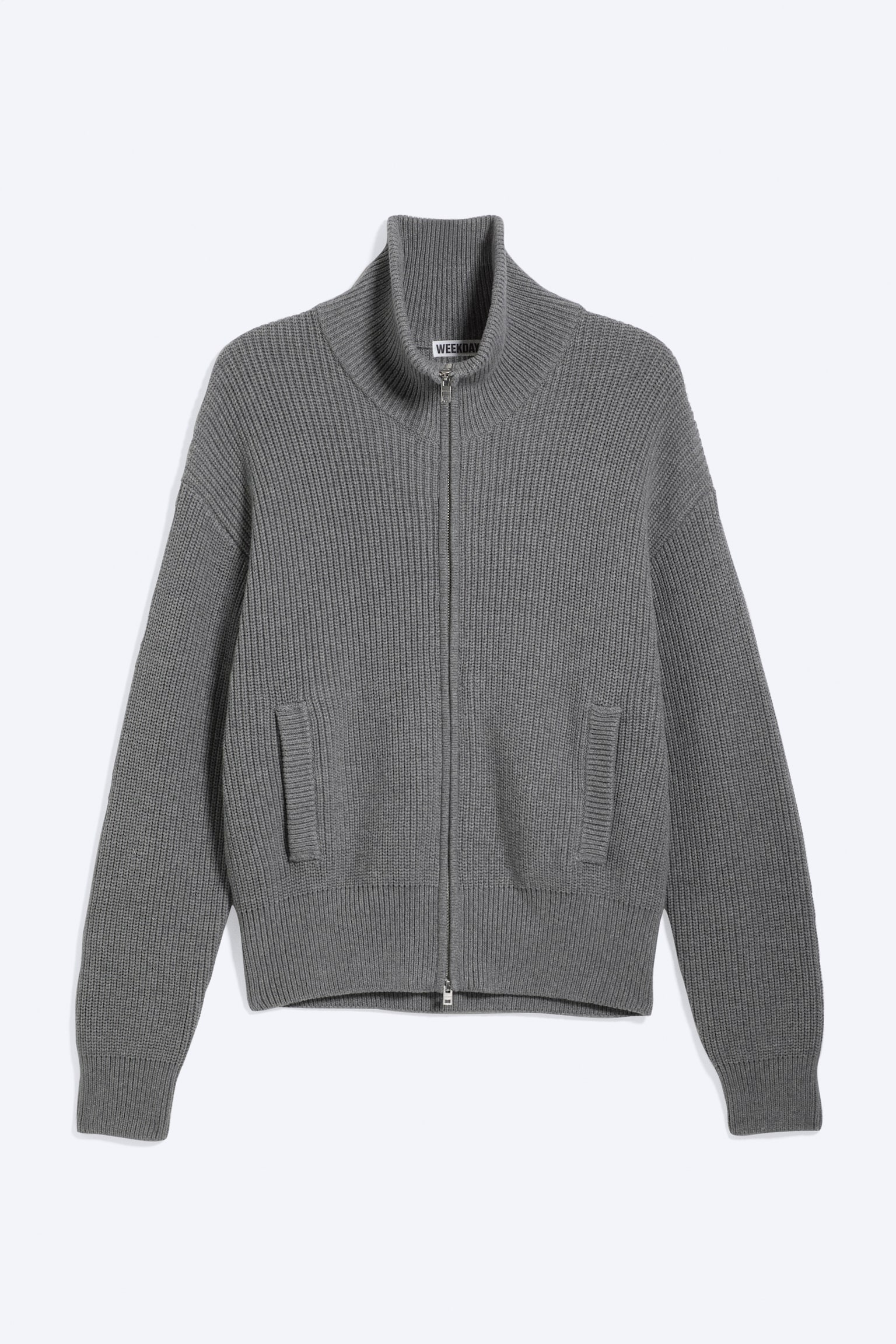Un cardigan en tricot gris est présenté de face, arborant une coupe décontractée avec un col montant et une fermeture éclair à double sens à l'avant. Il comprend des poches passepoilées latérales et est fini avec des poignets et un ourlet côtelés.