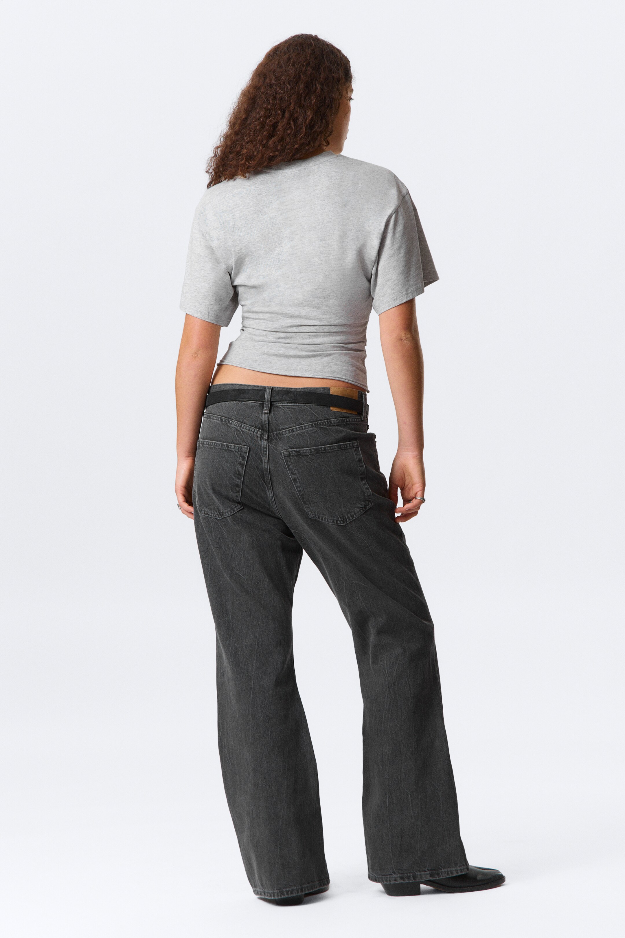 Agrandir l'image: Un t-shirt gris clair à manches courtes est montré de dos, avec un détail froncé au bas du dos qui crée un ourlet irrégulier et court. Des jeans gris foncé à jambes larges sont portés avec une ceinture noire visible, mettant en valeur les poches arrière et une coupe décontractée.