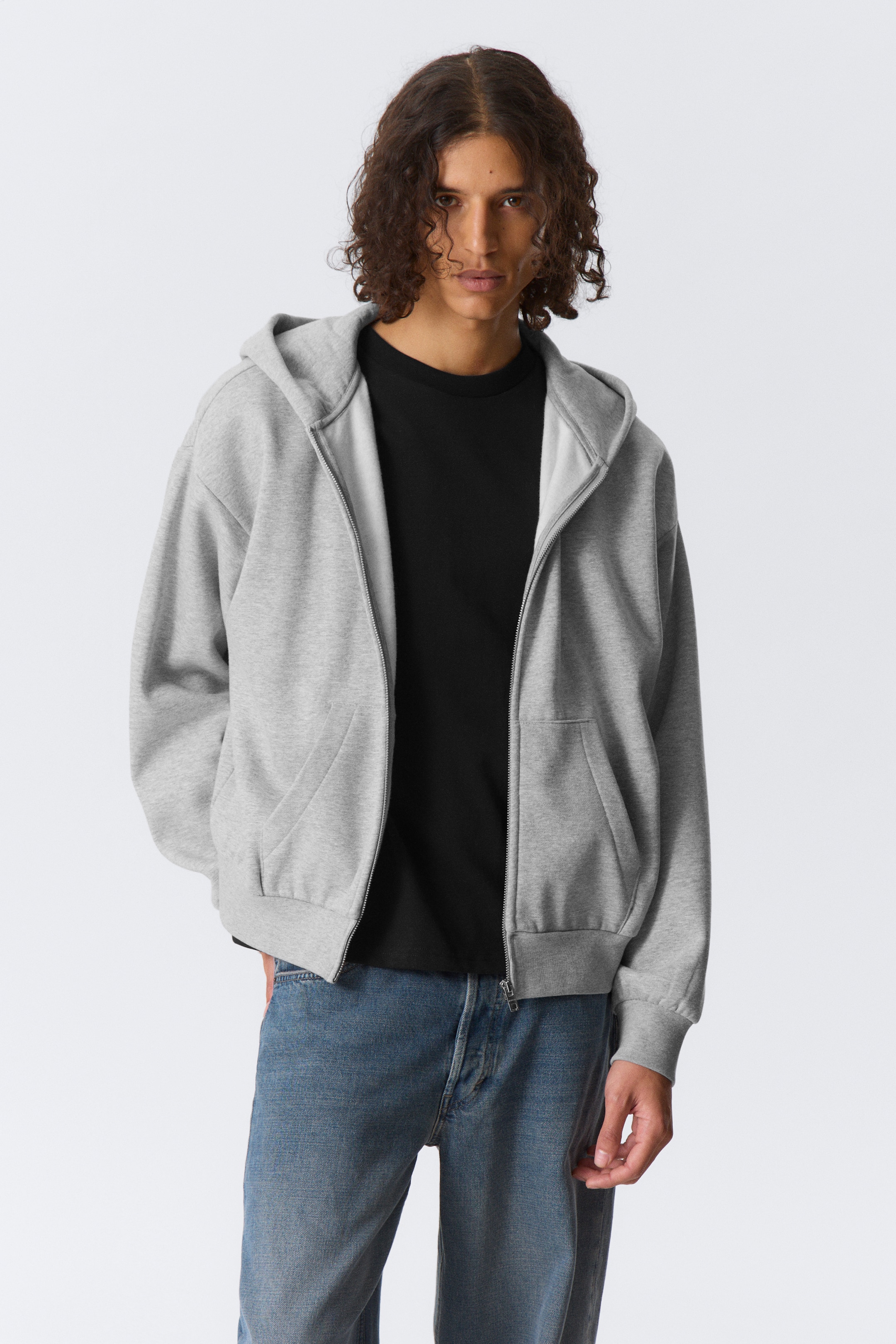 Grösseres Bild ansehen: Eine hellgraue Kapuzenjacke mit Reissverschluss wird nach vorne präsentiert, offen getragen über einem schwarzen T-Shirt. Sie verfügt über eine Kapuze, einen silberfarbenen Reissverschluss in voller Länge, zwei aufgesetzte Taschen vorne, gerippte Bündchen und einen gerippten Saum.