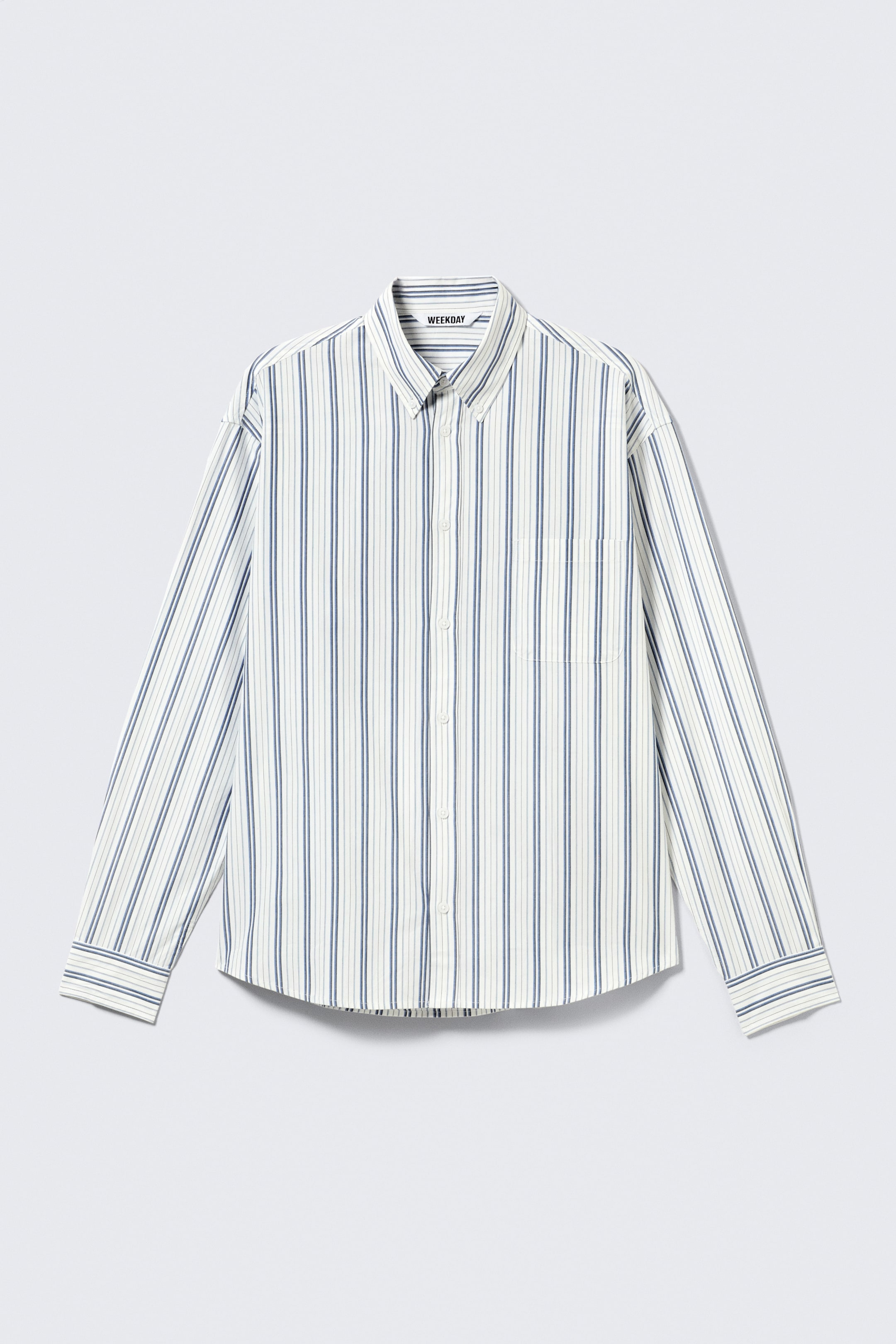 Ver imagen más grande: Una camisa de corte holgado y manga larga se muestra desde el FRENTE, con una base en color blanco roto y rayas verticales en azul marino y azul claro. Presenta un cuello con botones, hombros caídos, un bolsillo en el pecho, cierre de botones en la parte delantera, puños abotonados y un dobladillo curvado.