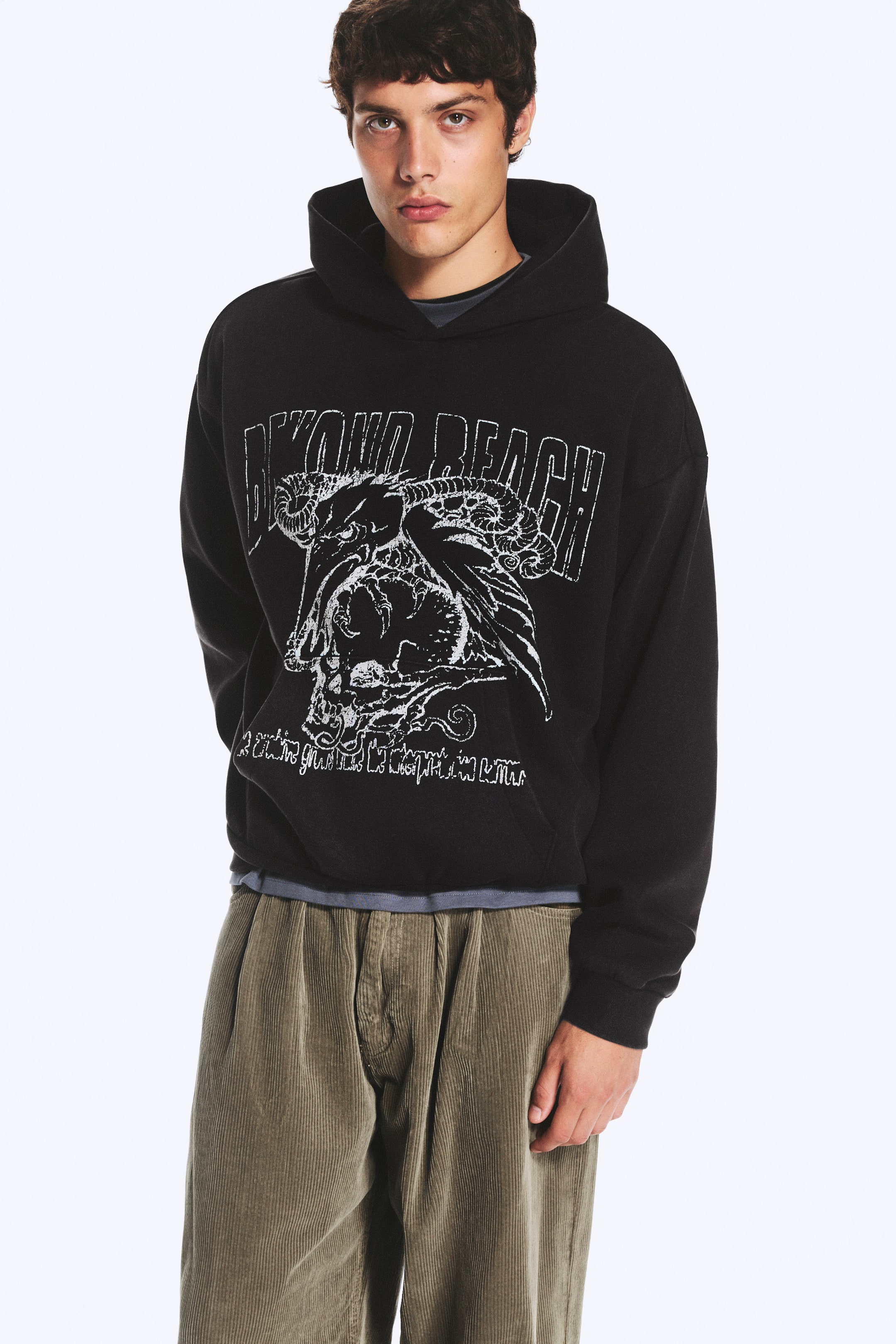 Grotere afbeelding bekijken: Een zwarte hoodie met losse pasvorm, voorzien van een grote witte grafische print over de borst, verlaagde schoudernaden en een ruime capuchon, wordt gedragen door het model. De hoodie is gelaagd over een grijs shirt dat zichtbaar is aan de zoom en kraag, en gecombineerd met olijfgroene ribfluwelen broek.