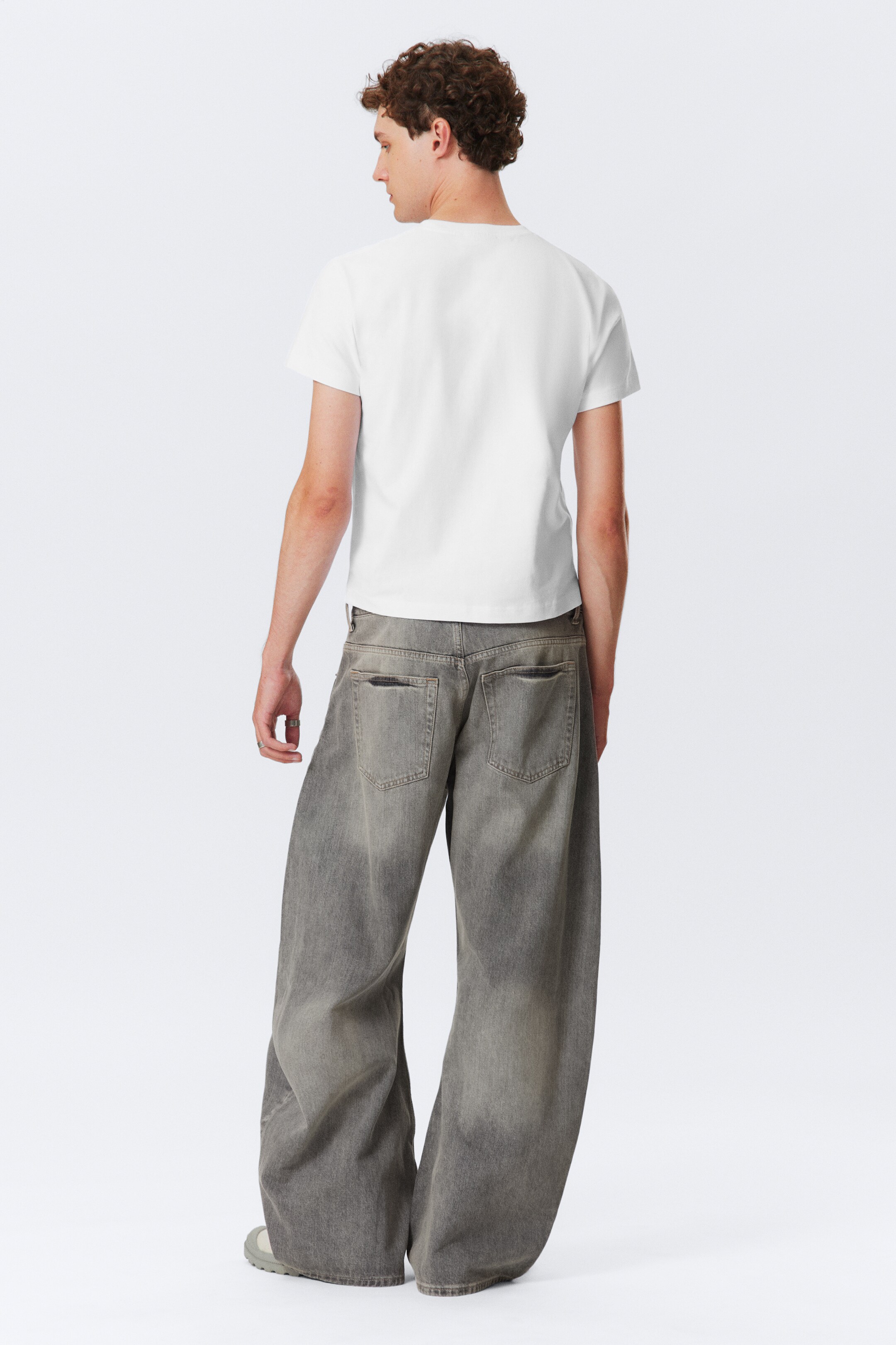 Agrandir l'image: Un t-shirt blanc à manches courtes et col rond est porté, vu de dos. Il est associé à un jean gris à jambes larges, qui présente un délavage et deux poches arrière visibles.