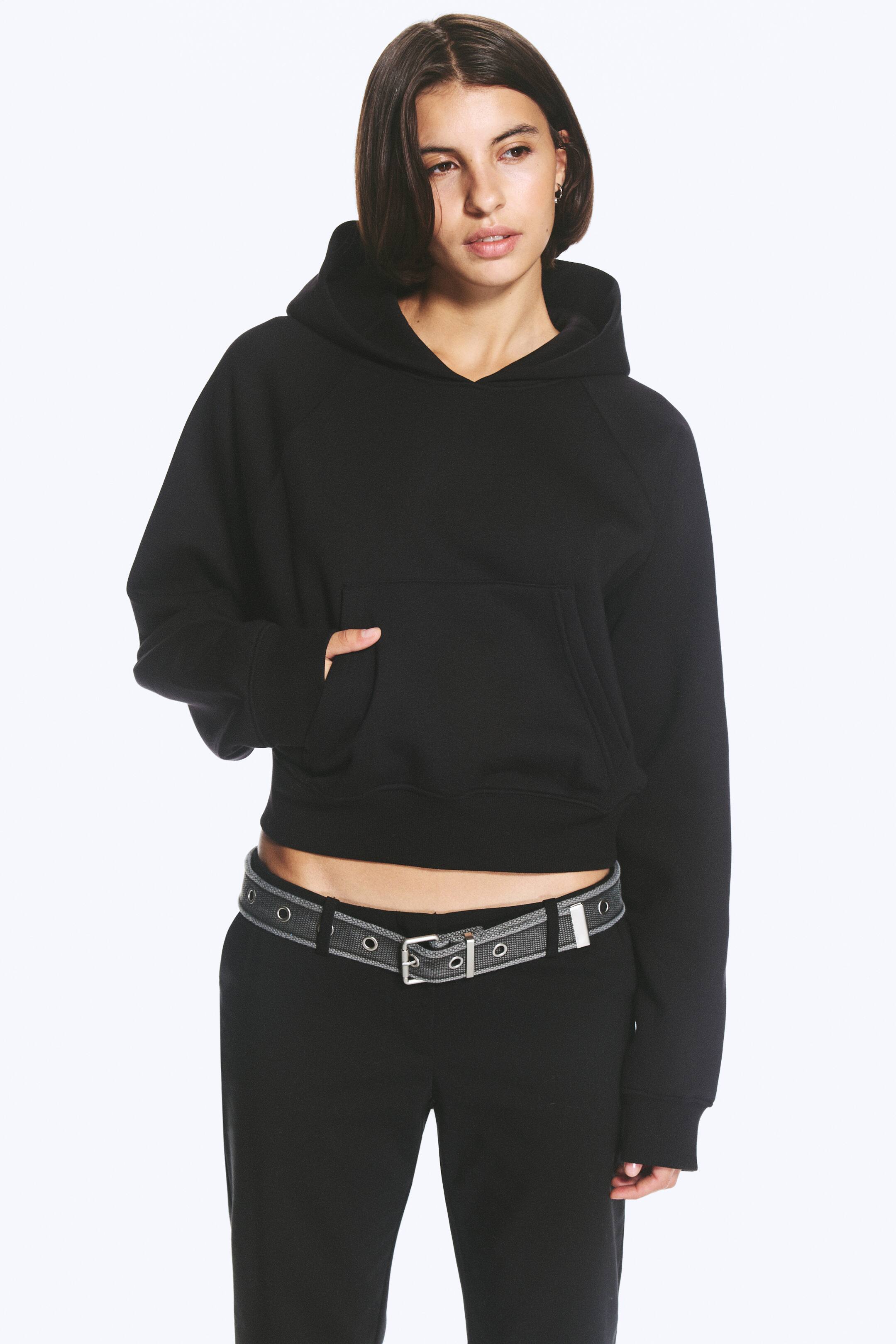 Grotere afbeelding bekijken: Een zwart sweatshirt met capuchon, voorzien van een ruime capuchon, raglanmouwen en een kangoeroezak, wordt gedragen door het model, dat één hand in de zak laat rusten. De sweatshirt met relaxed fit is afgewerkt met ribboorden aan de mouwen en zoom en gestyled met een zwarte broek en een grijze riem.