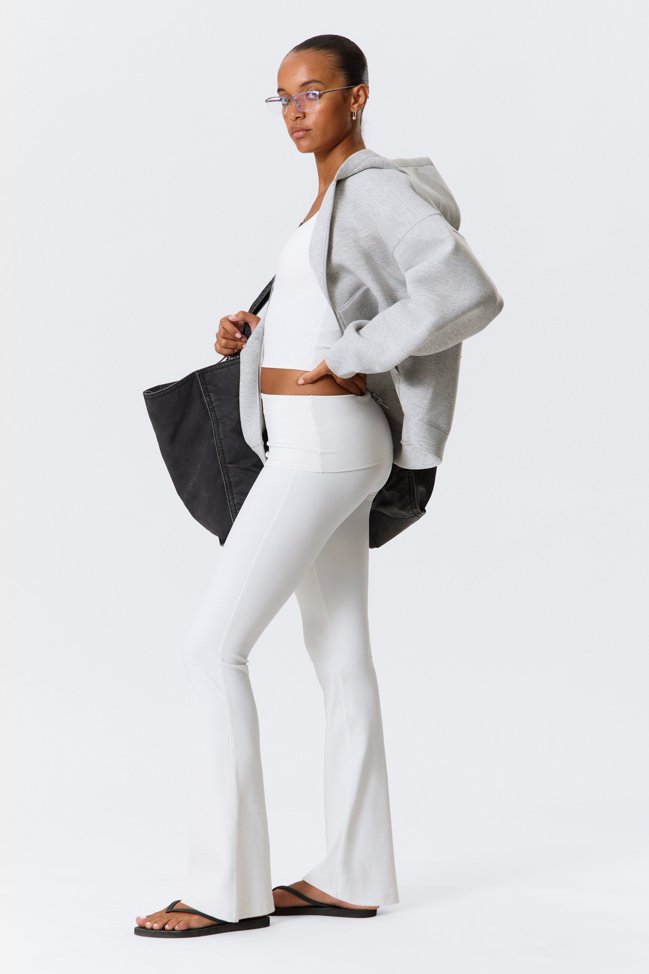 Agrandir l'image: Un pantalon blanc évasé à taille haute, ajusté à la cuisse et évasé à partir du genou, est porté avec un crop top blanc à encolure dégagée. Un sweat à capuche gris surdimensionné avec fermeture éclair est porté ouvert et drapé, tandis qu'un sac fourre-tout noir non structuré est porté et des tongs noires sont portées.